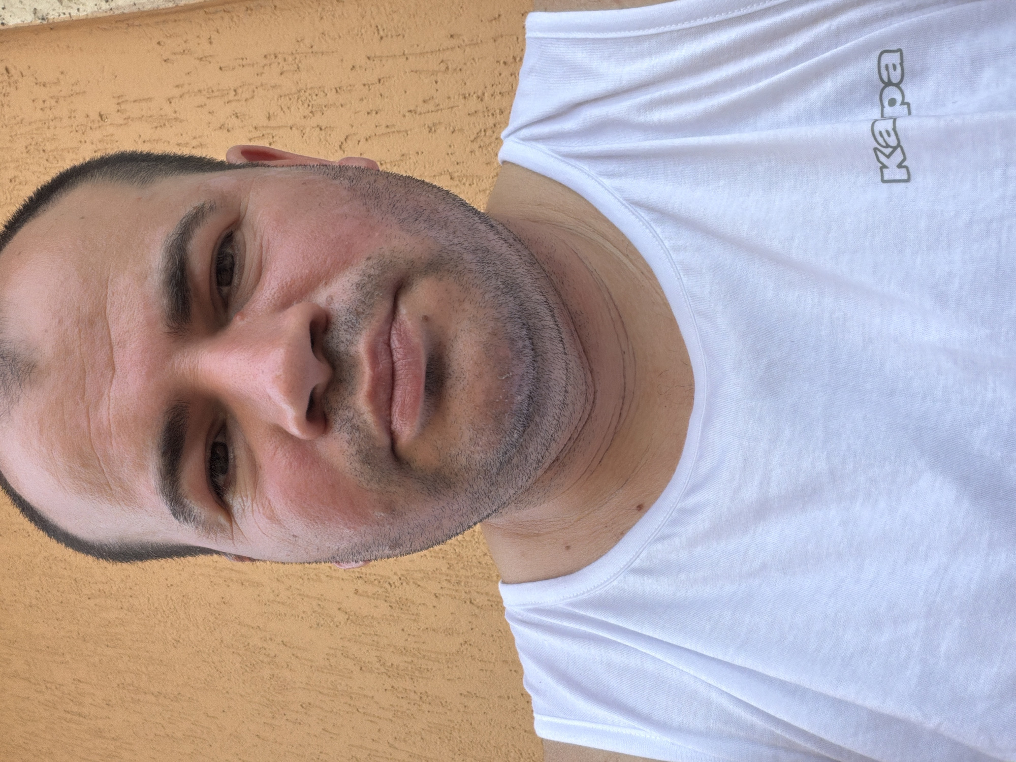 This is a close-up, head-and-shoulders shot of a man wearing a white t-shirt. The image is oriented vertically, with the man's head towards the left and his body extending to the right. He has short, dark hair and a stubbly beard. His skin appears slightly flushed, suggesting he might be outdoors or experiencing exertion. His eyes are dark and looking slightly off-camera. The t-shirt has a printed logo on the upper right chest area, which appears to read "Kapa" in a vertical, gray font. The background is a textured, orange-brown surface, possibly a wall or stucco, consistent with outdoor settings in Italy. The lighting suggests it is daytime, possibly sunny, given the bright illumination on the man's face and t-shirt. No specific event is depicted, but the selfie-style framing and casual attire suggest a personal moment or a casual portrait. The overall impression is one of a candid, informal portrait captured outdoors. The location context provided is Piacenza, Italy.