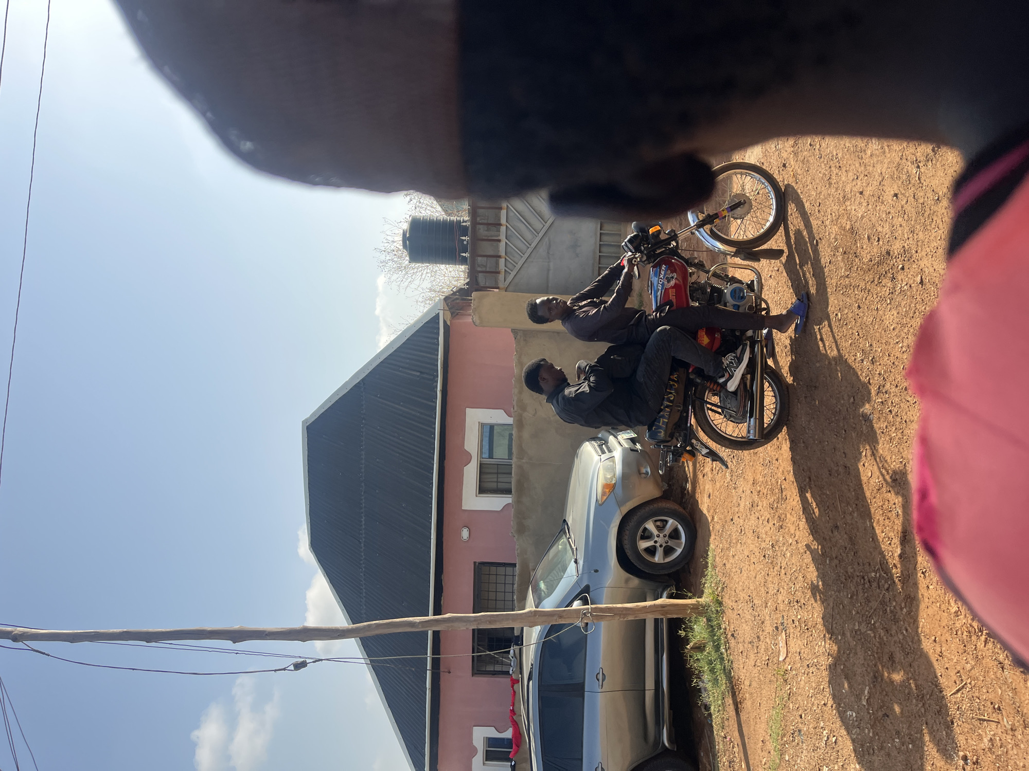 Two men are on a red motorcycle parked on a dirt road next to a silver car. A house with a black roof and pink walls stands in the background, near a water tank. The scene appears to be in a residential area in Abuja, Nigeria, with utility poles visible in the background. The image appears to be taken from a low angle, with a portion of the photographer's head visible.