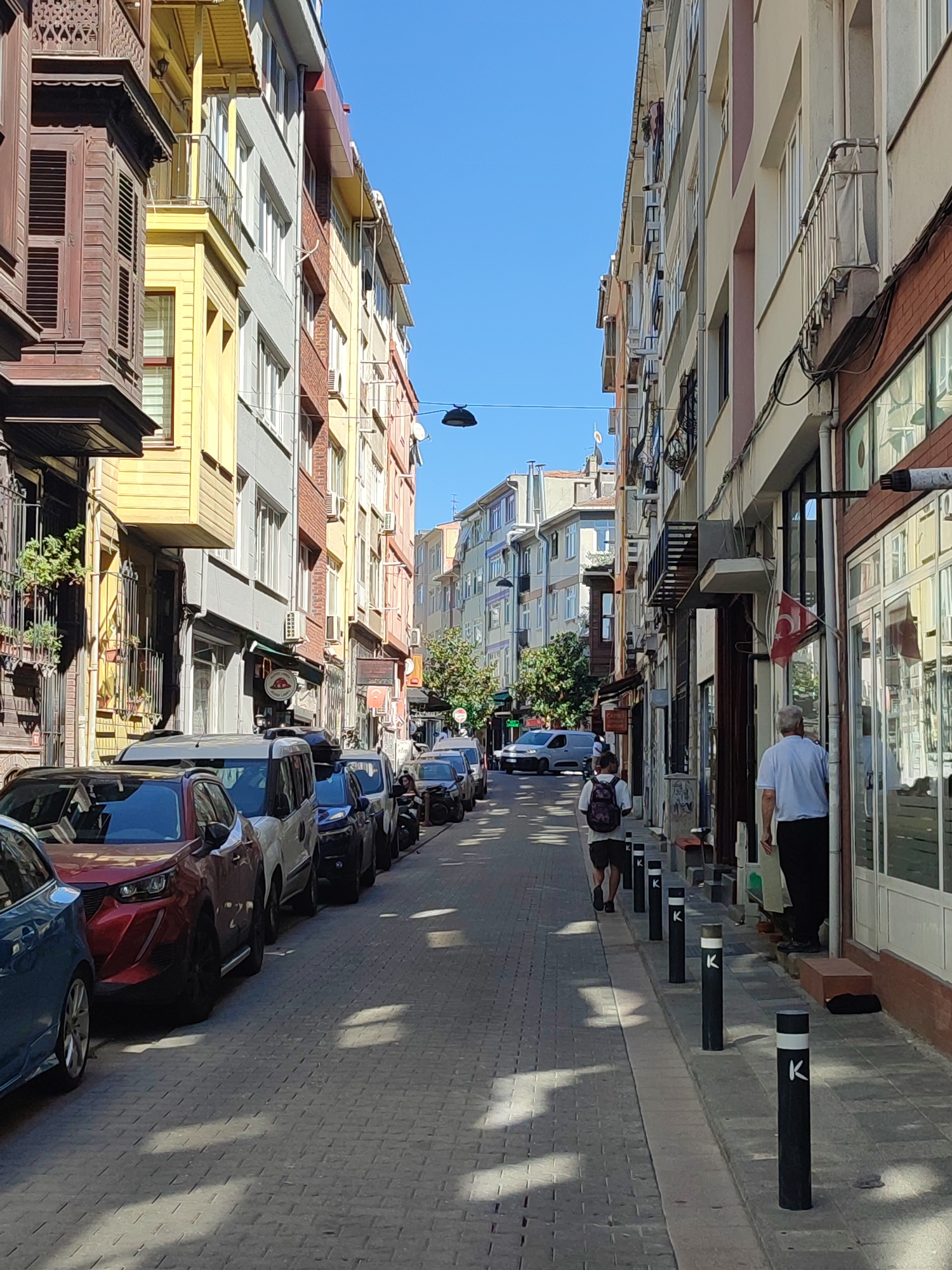 The image captures a narrow, cobblestone street in Kadıköy, Turkey, lined with multi-story buildings on both sides. The architecture displays a mix of traditional wooden elements, particularly on the left side, and more modern construction with balconies and varying colored facades. The street is filled with parked cars, with the closest vehicles being red and blue sedans. Two individuals are visible: one person with a backpack walking away from the camera down the street, and another person, a man in a white shirt and dark pants, standing near a shop entrance on the right. The sky is clear and blue, and sunlight casts distinct shadows on the street and buildings, indicating it is daytime. Shops and businesses appear to be situated on the ground floors of the buildings, with windows and signage visible. There are also street lamps and bollards present along the street.