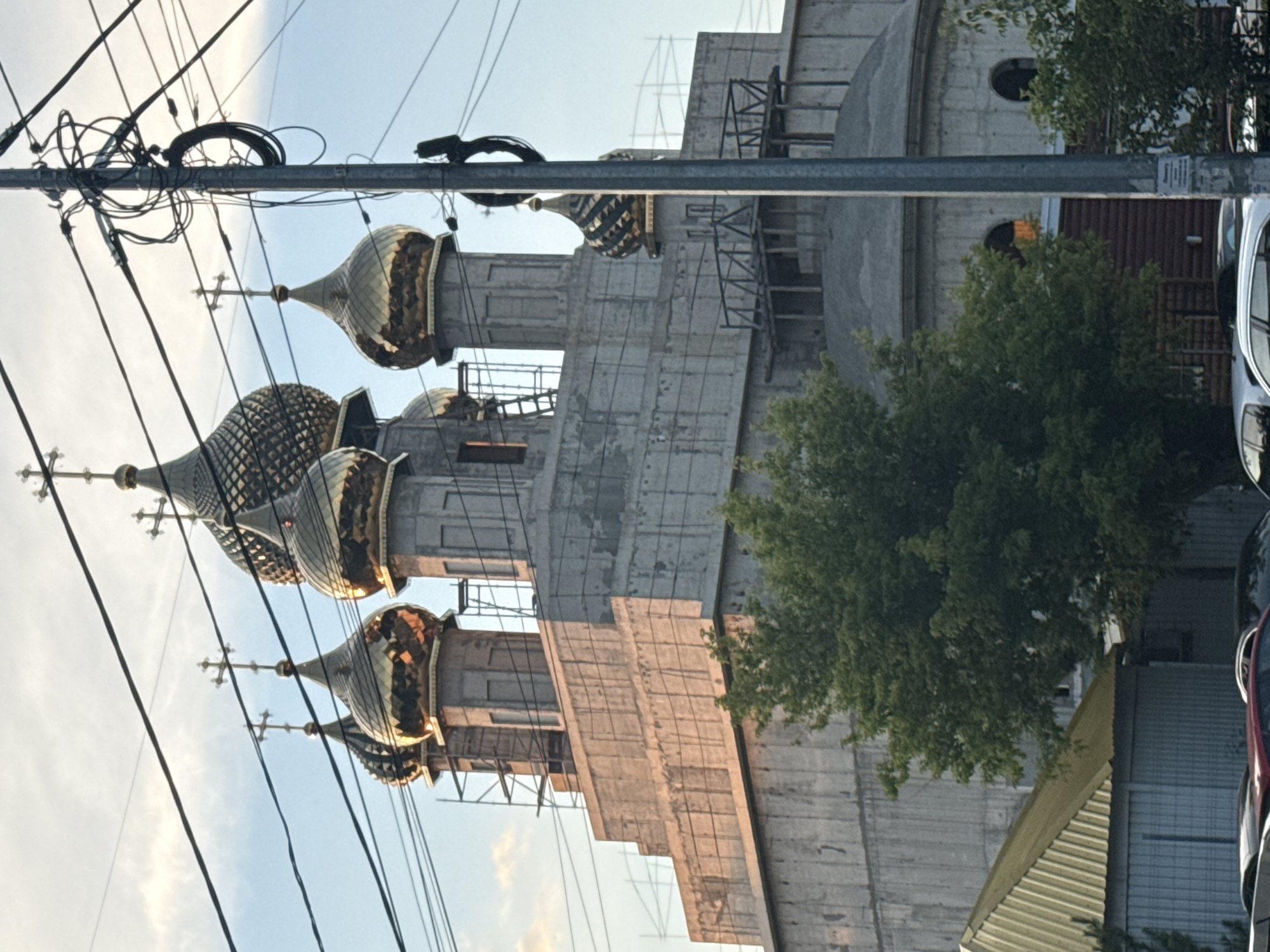 A construction of an Orthodox church is captured in this medium shot. The church has several golden domes with crosses on top, and scaffolding is visible around the structure. The building is made of concrete and appears to be in the early stages of completion. Power lines and a utility pole are in the foreground, partially obscuring the church. A lush green tree stands to the left of the church, and a yellow metal roof of a smaller structure is visible in the lower left corner. Several cars are parked in front of the buildings. The sky is a clear blue with some scattered clouds, and the lighting suggests it is late afternoon or early evening.