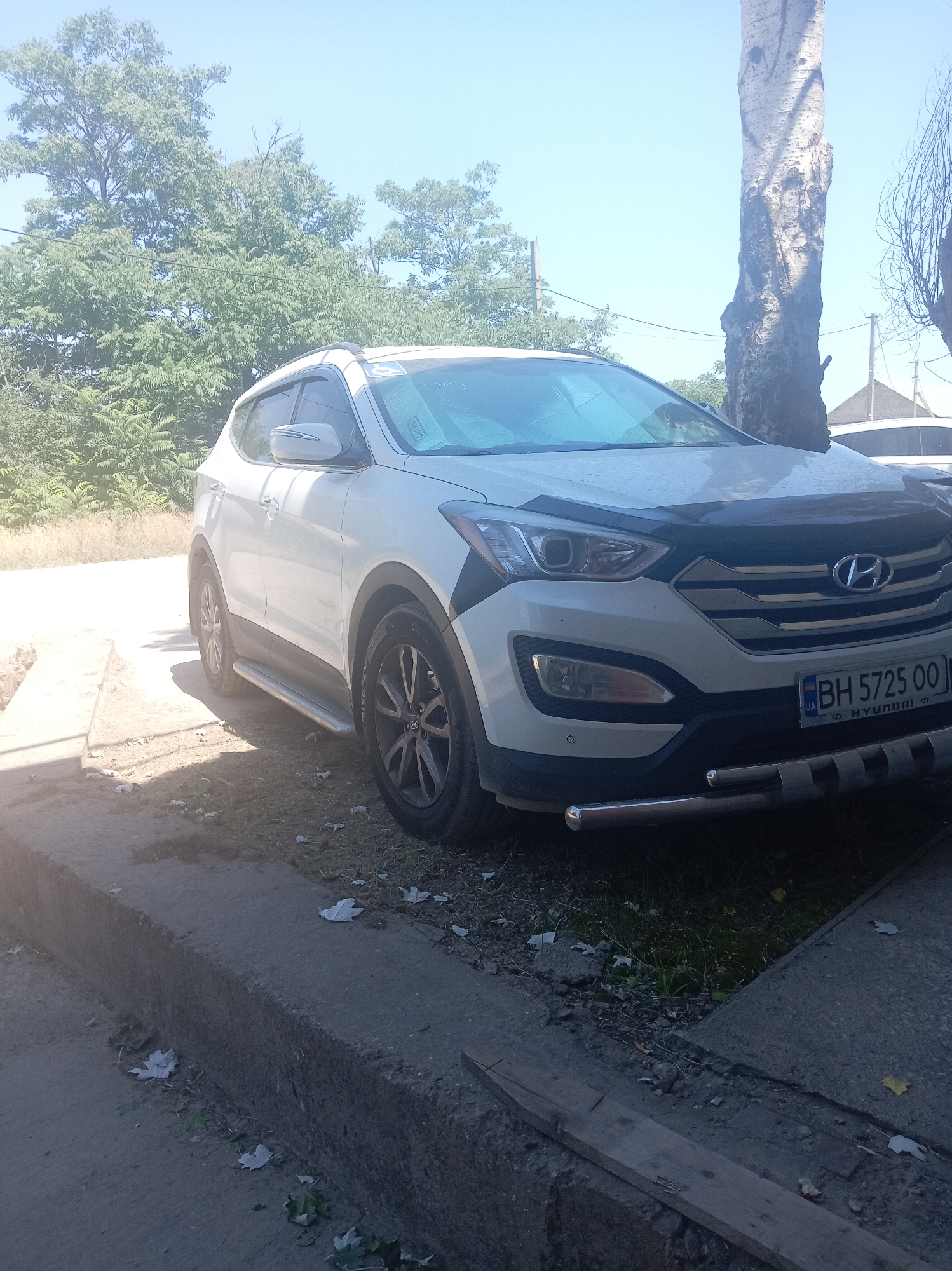 The image shows a white Hyundai Santa Fe parked on a street in Velykodolynske, Ukraine. The car is positioned at an angle, with its front facing the viewer. The scene appears to be outdoors, with a bright, sunny sky suggesting daytime. Lush green trees form the background, with a prominent, tall, weathered tree trunk to the right of the car, partially obscuring a distant building. The foreground shows a concrete curb and street surface, with scattered leaves and debris. The car has a black hood deflector and a chrome bull bar on the front bumper. The license plate is visible, displaying "BH 5725 00 UA". The overall impression is that of a vehicle parked in a semi-rural or suburban environment on a clear day. There are no people visible in the image.