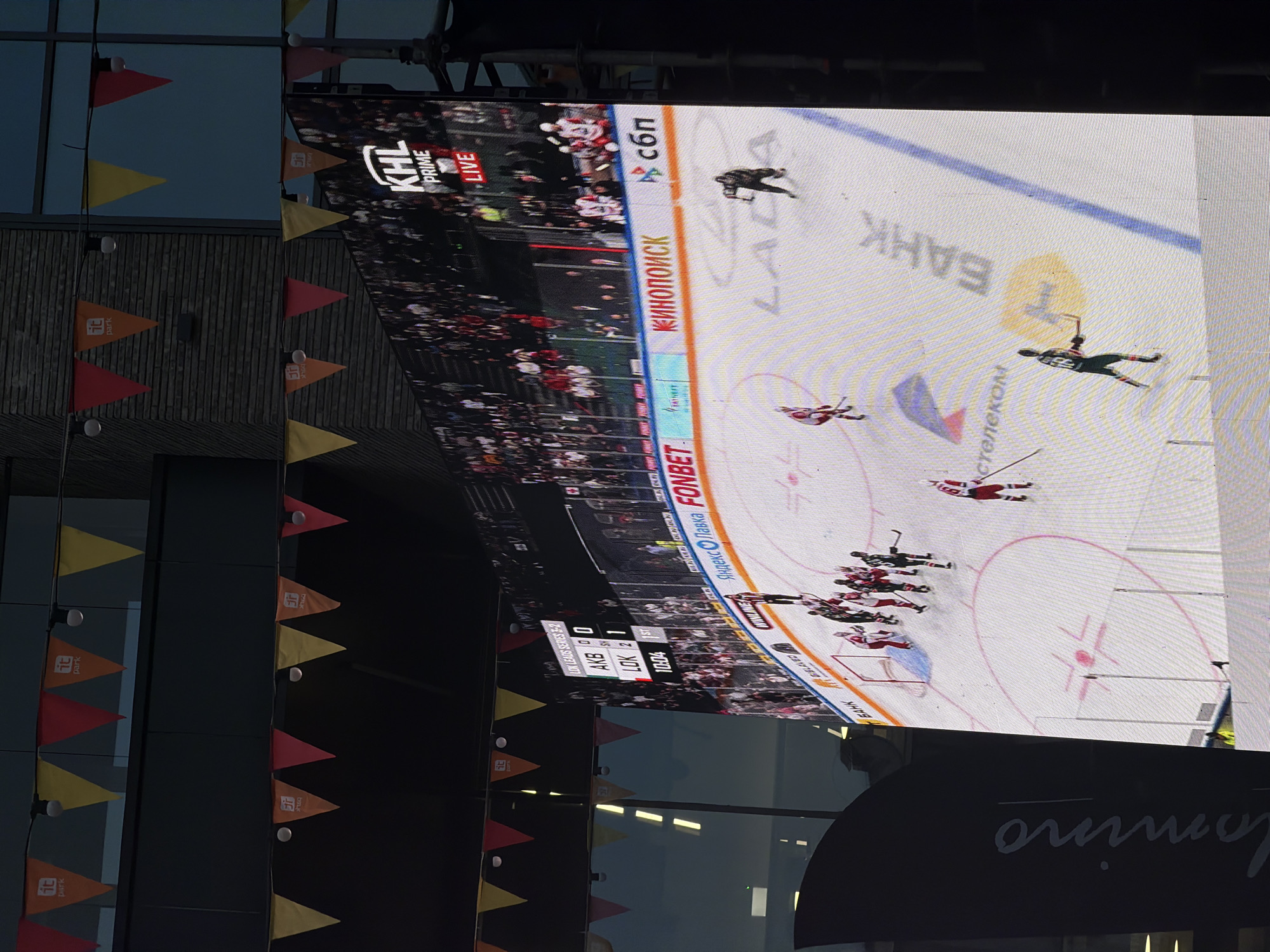 The image shows a large outdoor display in Kazan, Russia, likely at IT Park, broadcasting a live KHL Prime hockey game between AK Bars Kazan (AKB) and Lokomotiv Yaroslavl (LOK). The score is AKB 0, LOK 2 in the first period, with the time at 10:04. Players are visible on the ice. Several sponsor logos surround the ice rink, including Fonbet, Yandex Lavka, Kinopoisk, and Sberbank. Triangular pennants decorate the building, which features the IT Park logo. The word "lomino" is also visible on the bottom right.