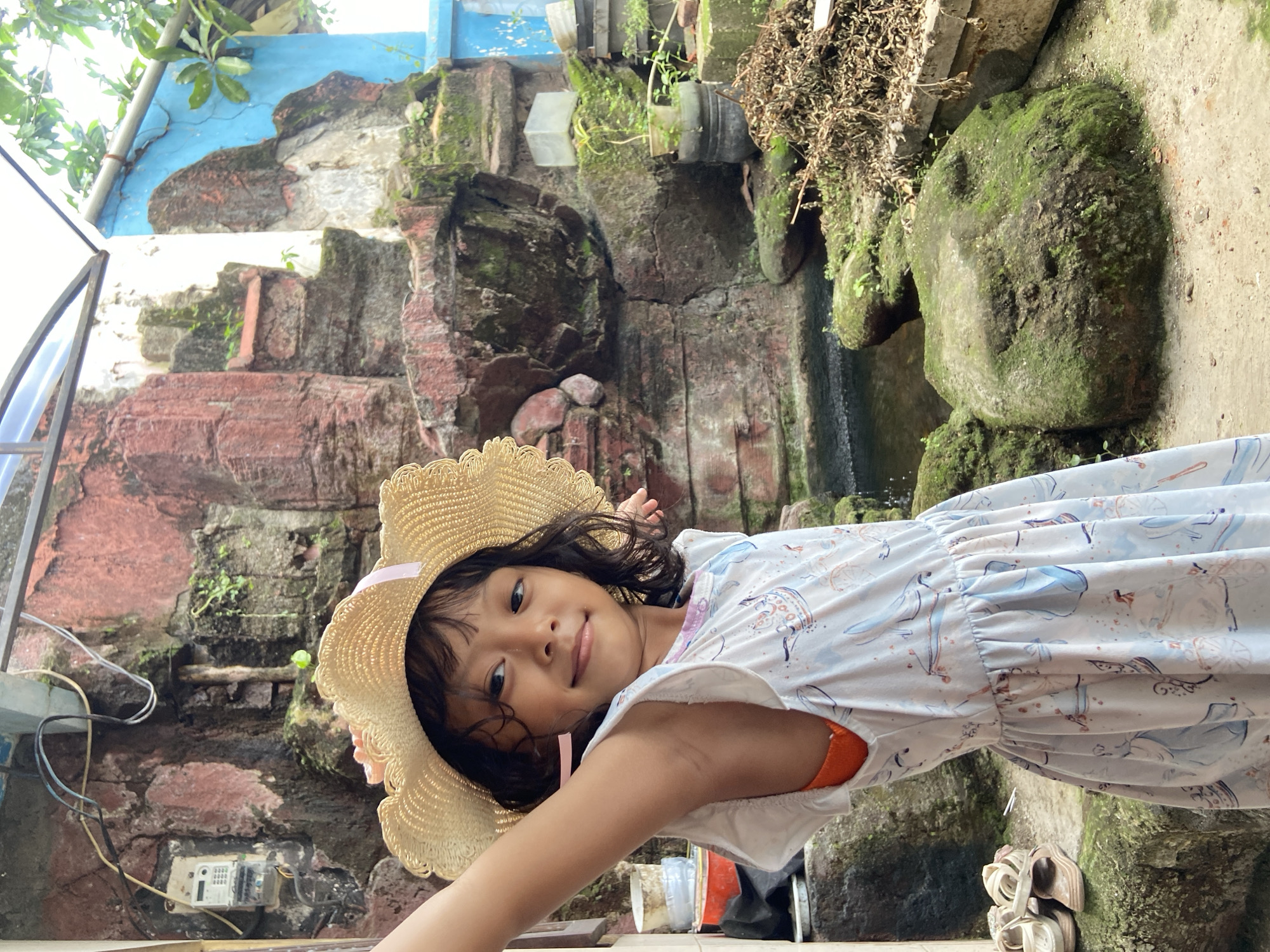 A young girl with dark hair wearing a straw hat and a light-colored patterned dress is the primary subject. She is looking towards the camera with a slight smile. The background features a complex, multi-tiered structure, possibly a water feature or garden design, constructed from brick or stone. It appears aged, with moss and vegetation growing on it. The presence of electrical wires and a meter on the lower left suggests an urban or suburban environment. The image appears to be taken in daylight, with natural light illuminating the scene. Further back in the image, there are additional plants and some signs of residential construction (e.g., blue paint on a wall), indicating a location likely in the city of Gelam, Indonesia.