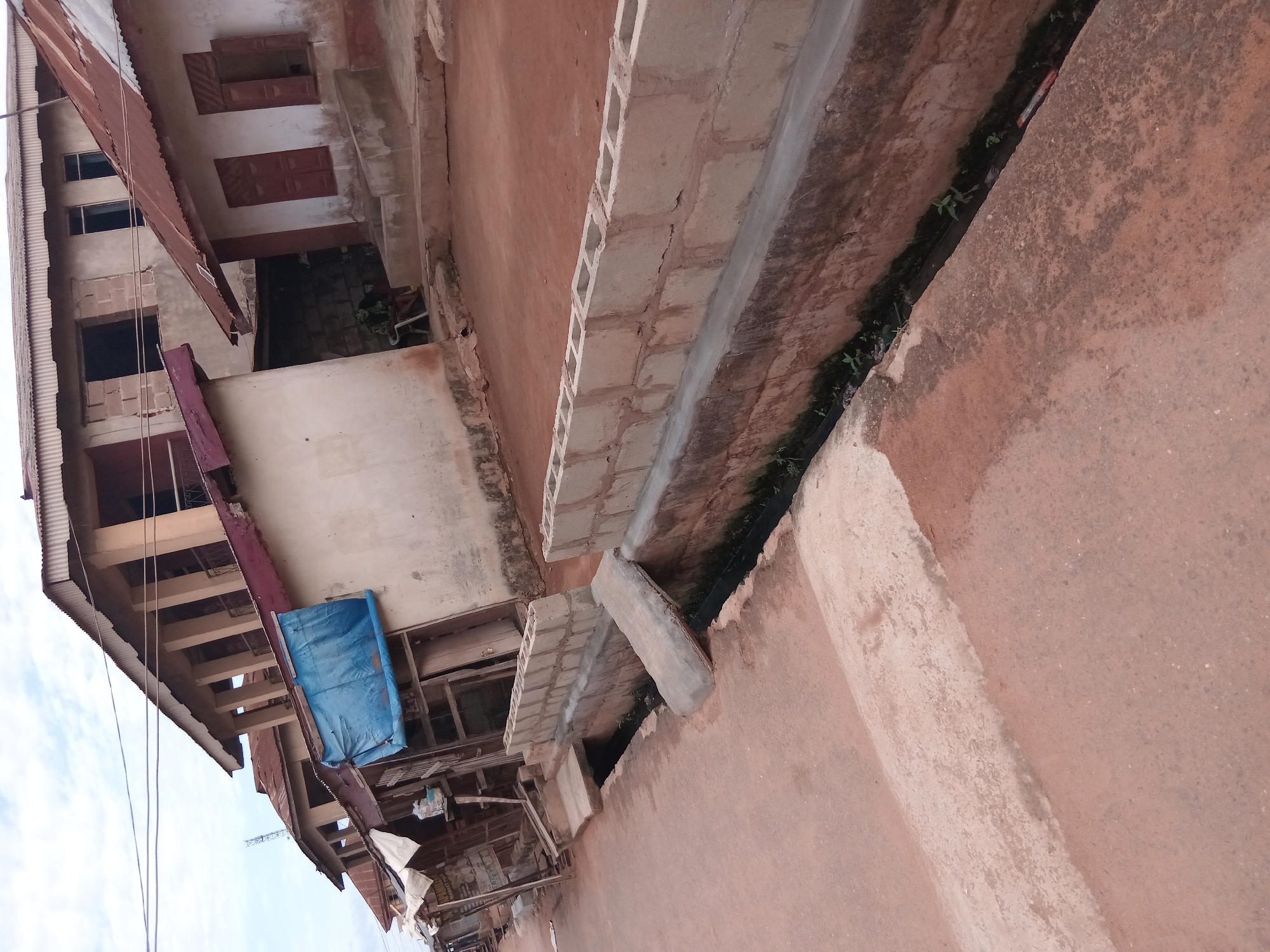 The image depicts a street scene in Shagamu, Nigeria. The perspective is tilted. Buildings line the street, constructed of concrete block and with corrugated metal roofs. One building is three stories tall. The street is paved, showing wear and discoloration. There are weeds growing in the cracks and seams between the paving slabs. A blue tarp is visible on one of the buildings. The scene appears to be daytime, with a blue sky visible. There are no people visible.