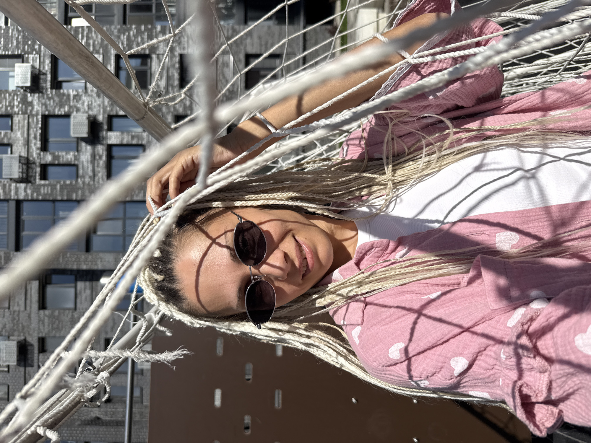 In Rusanovo, Russia, a woman with blonde braids and sunglasses is posing for a photo. She is wearing a pink shirt with white hearts and a white t-shirt underneath. The background shows a modern building with many windows and air conditioning units. The woman is partially obscured by a white net, possibly part of a sports facility. Sunlight casts shadows on her face and clothing.