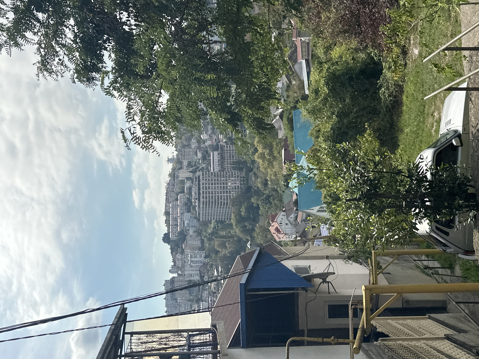 The image depicts a panoramic view of a city nestled on a hillside, likely in Sochi, Russia, given the architecture and vegetation. The foreground features a portion of a white building with a dark blue awning and brown tiled roof, accompanied by yellow metal scaffolding and a decorative fence. A white car is parked on a grassy incline to the right, partially obscured by greenery. In the mid-ground, several houses with various roof colors, including teal and red, are visible amongst dense foliage. The background reveals a densely populated urban area with numerous multi-story buildings of varying architectural styles, stretching across the undulating terrain towards the horizon under a partly cloudy sky. The overall scene suggests a residential district with a mix of older and more modern structures, characteristic of a coastal city with hilly topography. There are no people visible in the image.
