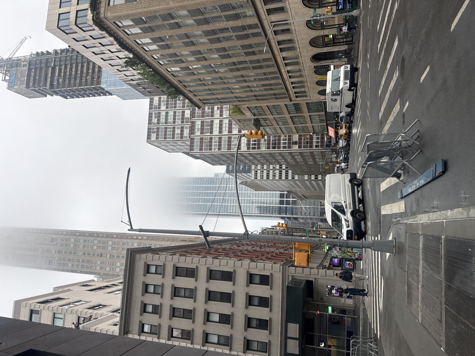 The image shows a street scene in New York City on a foggy or overcast day. The perspective is from a street corner, looking down a wide city street lined with tall buildings.
In the foreground, there is a crosswalk with white stripes on the wet asphalt. Several metal barricades are positioned on the sidewalk and street. A manhole cover is visible in the crosswalk. To the left, a dark grey building occupies the edge of the frame, and next to it is a lighter colored building with numerous windows. Further down the street, to the left, is a white cargo van.
The middle ground features a street with several cars and a white postal service truck. Traffic lights are visible, with one showing yellow. Pedestrians are crossing the street.
In the background, multiple skyscrapers dominate the skyline. One very tall building in the center is partially obscured by fog. To the right, a building under construction with a crane on top is visible, along with other office buildings. The sky is grey and overcast, contributing to the diffused lighting. The buildings display architectural styles typical of urban environments, with many windows and varying facade materials. The street appears to be a busy commercial or financial district.