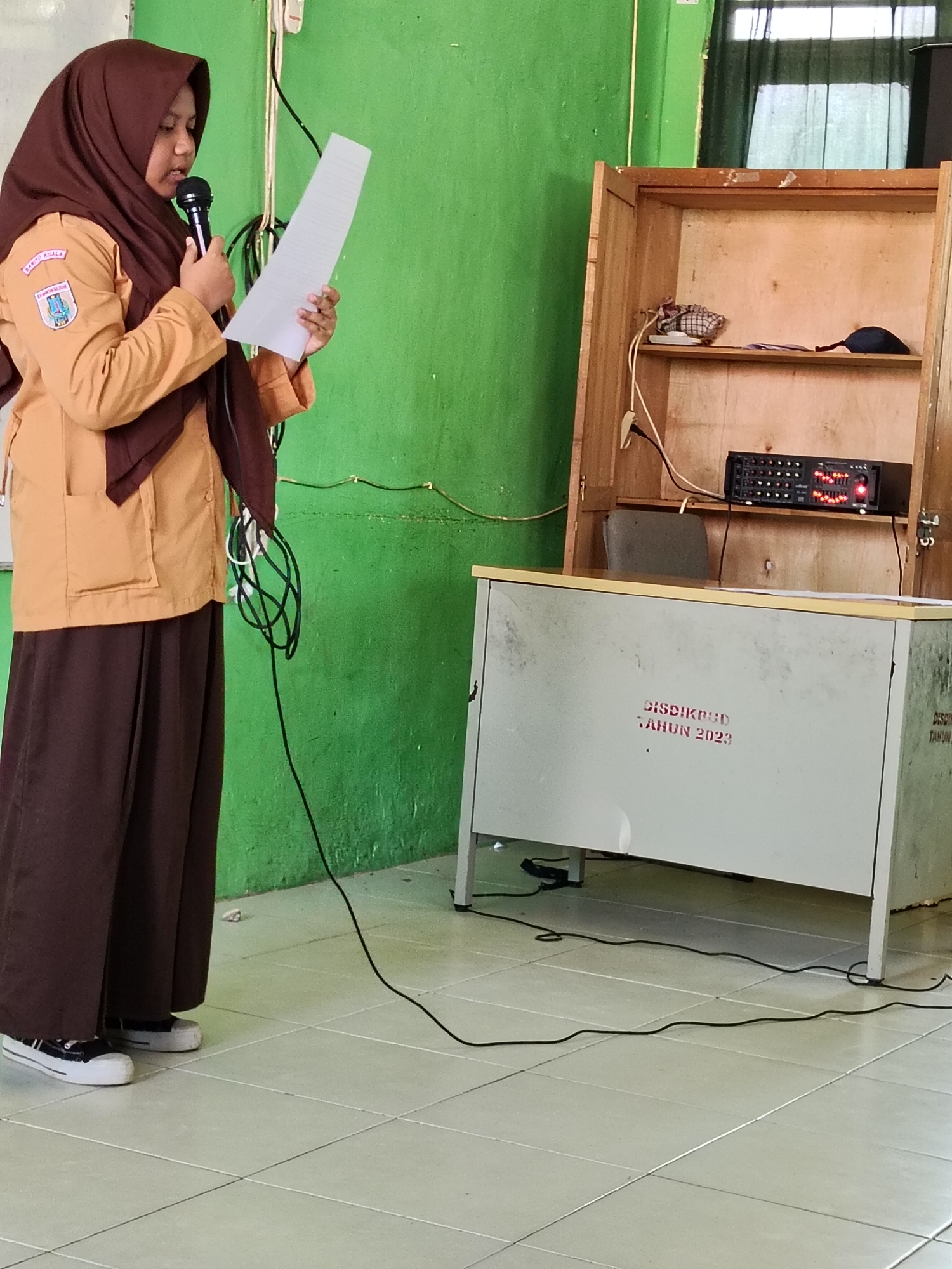 A young girl wearing a brown hijab and a tan school uniform stands in front of a green wall, holding a microphone and a piece of paper. She is speaking into the microphone, appearing to be giving a presentation or speech. To her right, there is a wooden cabinet with shelves, containing a few items. In front of the cabinet, there is a white desk with some writing on it. Electrical cords are visible on the floor and running along the wall. The floor is tiled. The image appears to be taken indoors, possibly in a school or community center in Lori, Indonesia.