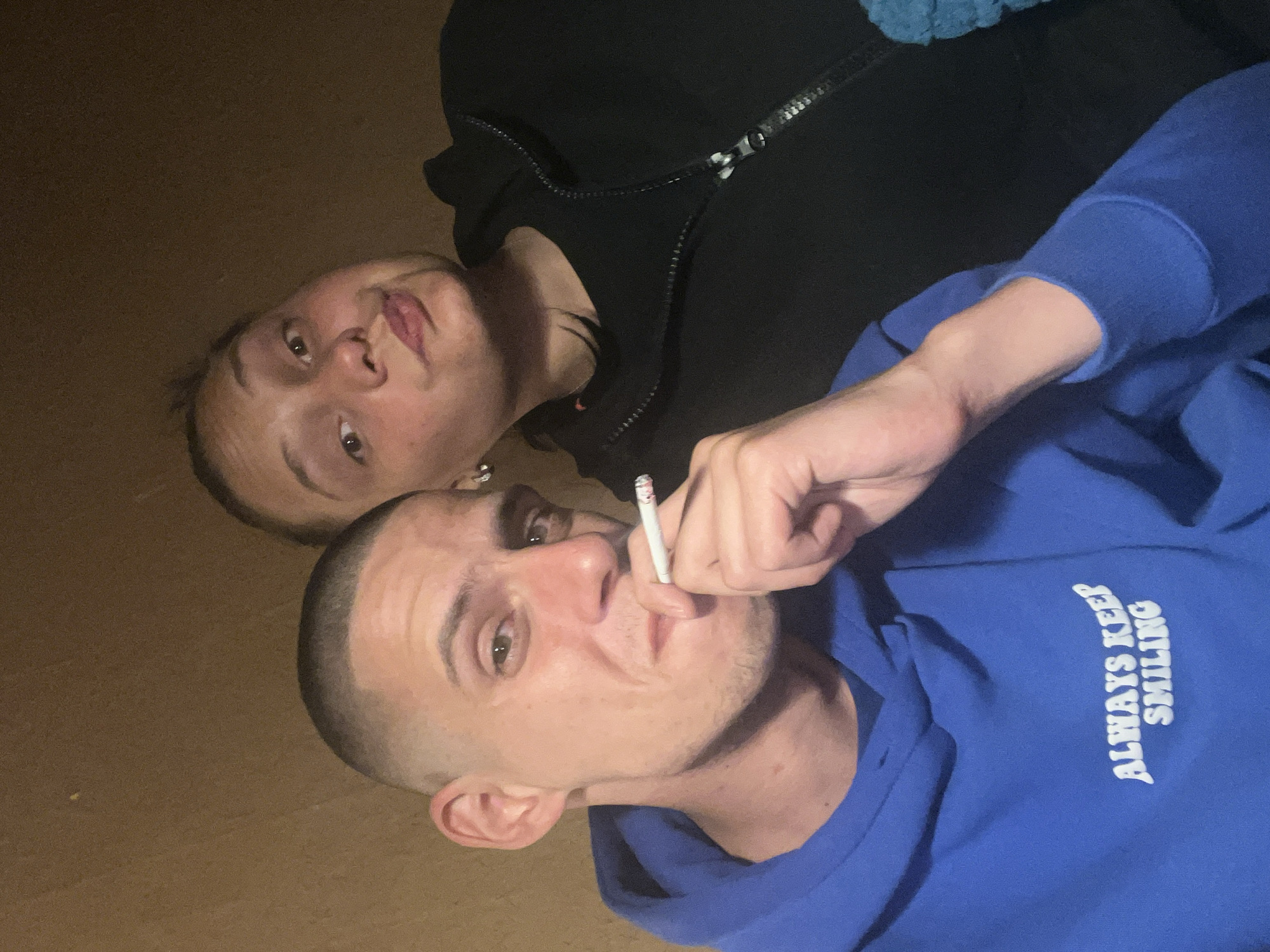 Two individuals are lying down on a brown surface, possibly a floor, indoors. One person is wearing a black hooded jacket, and the other is wearing a blue hooded sweatshirt with the words "ALWAYS KEEP SMILING" printed on the right side. The person in the blue sweatshirt is holding a lit cigarette in their right hand. There is no visual information that suggests the location is Sofia, Bulgaria.