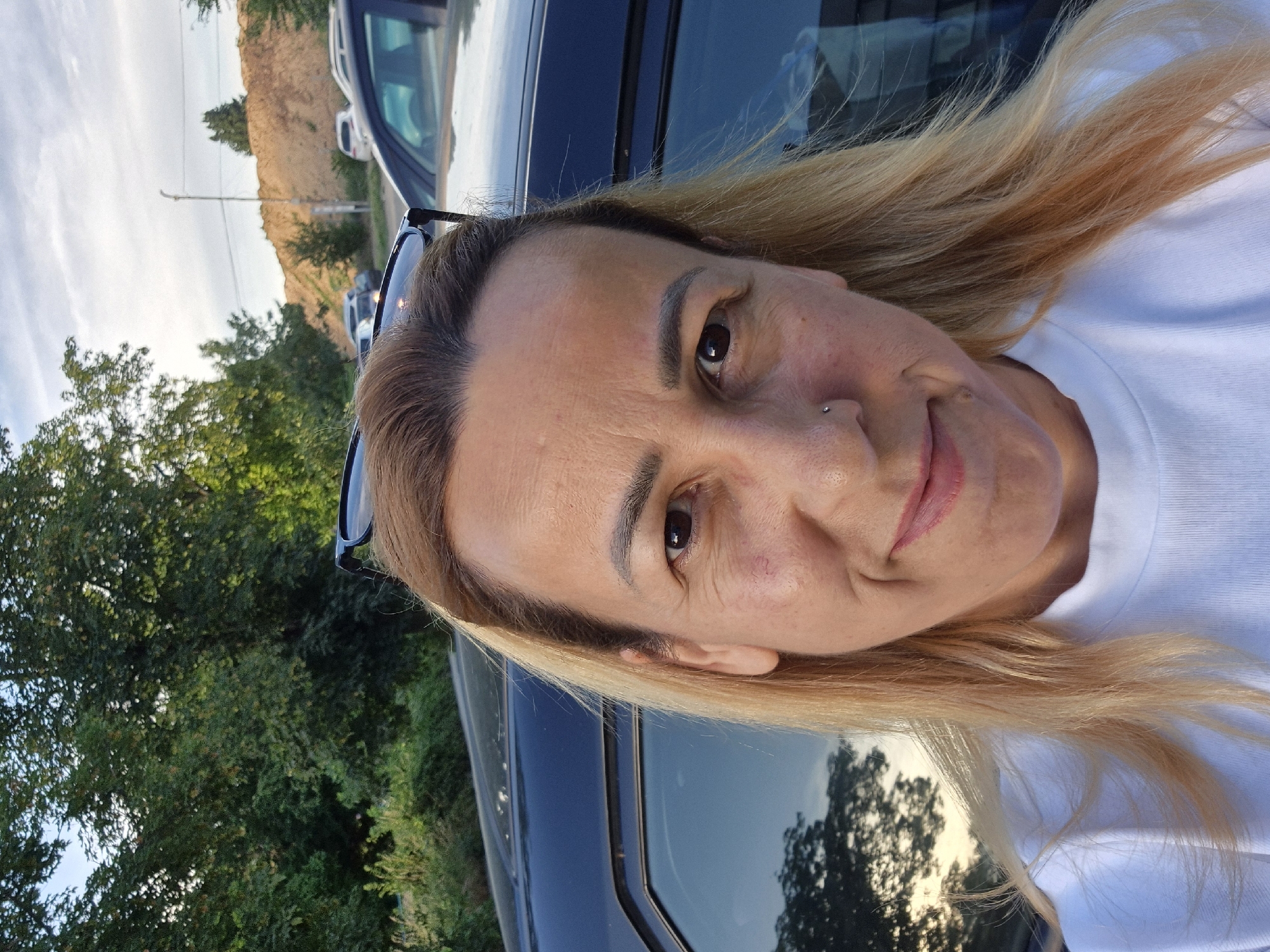 A woman with blonde hair is taking a selfie outdoors. She is wearing a white t-shirt and sunglasses are perched on top of her head. Behind her, a dark-colored car is visible, along with trees and a dirt mound. The sky is cloudy.