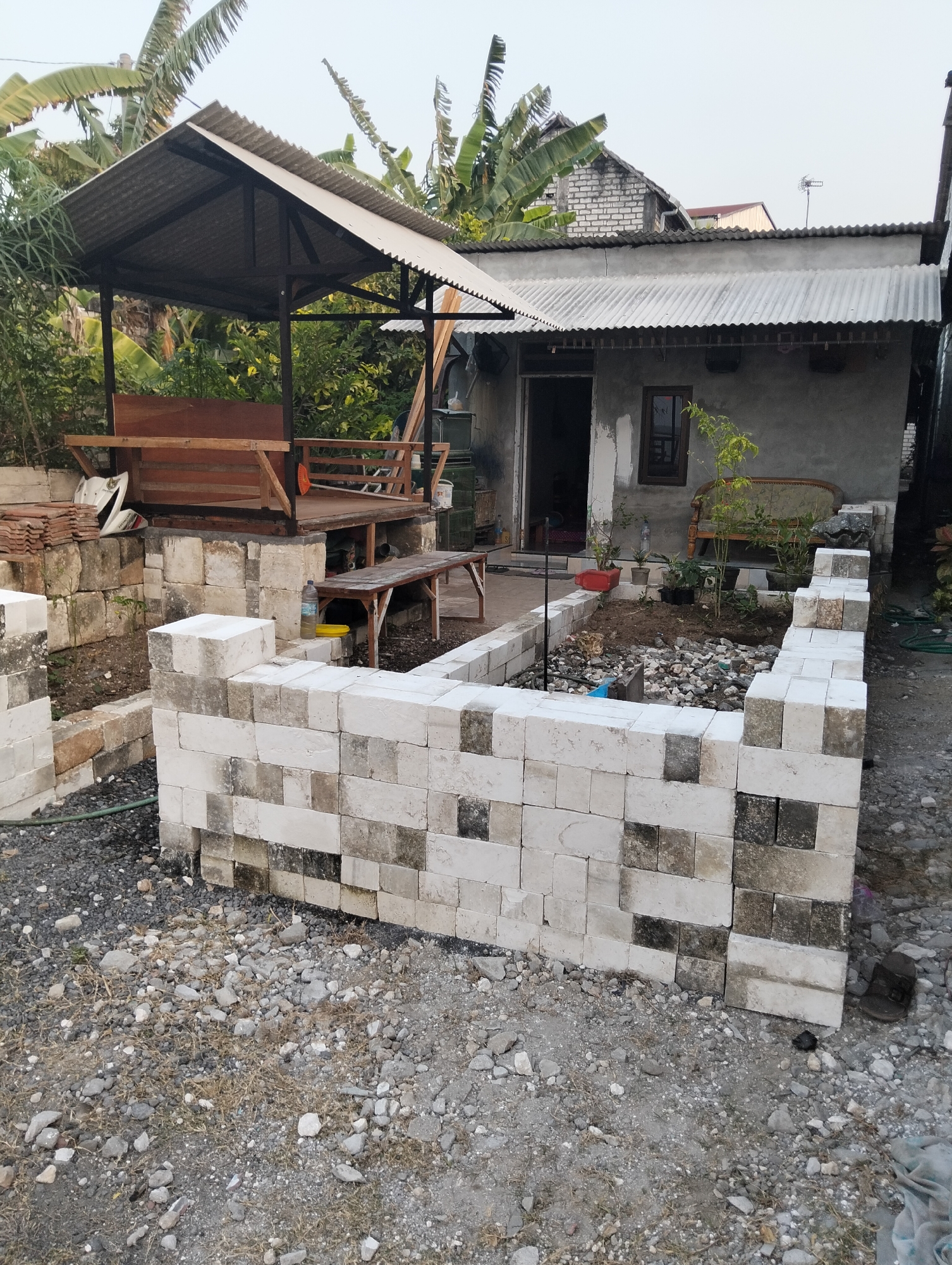 The image depicts an outdoor scene in what appears to be a residential area in Paciran, Indonesia. In the foreground, a low wall constructed from white and greyish-white bricks is partially built, forming a rectangular enclosure. The ground around it is covered in gravel and small stones. Behind this wall, to the left, is a wooden structure with a corrugated metal roof, resembling a gazebo or open-sided shelter, with wooden benches inside. Further back, a simple house with a gray concrete exterior and a corrugated metal roof is visible. An open doorway leads into the house. In front of the house, there is a garden area with plants and a sofa with a patterned cushion. To the right of the house, a narrow alleyway or passage is partially visible. The overall impression is of an unfinished or work-in-progress outdoor space. There are no people visible in the image.