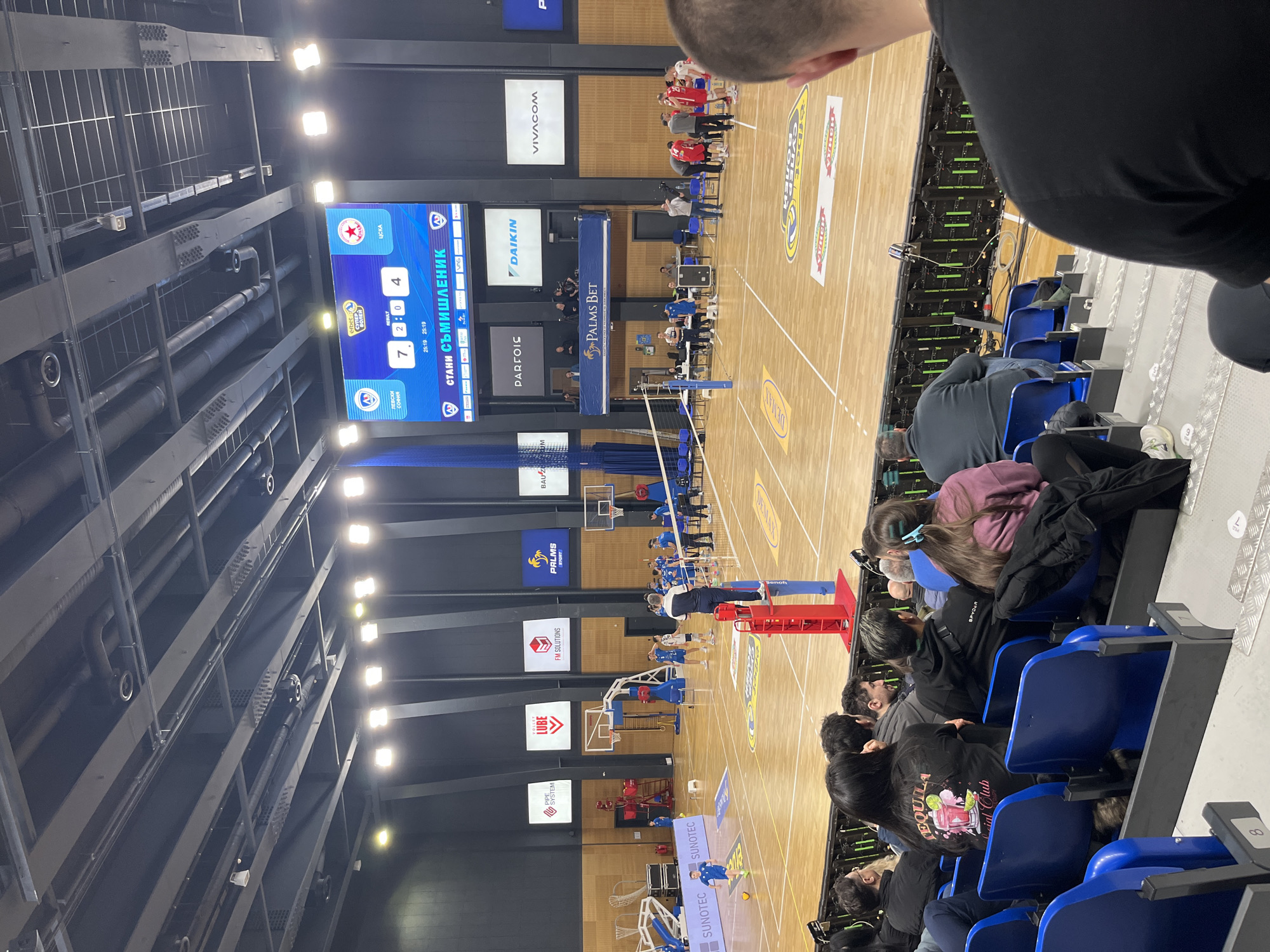 The image shows a volleyball match being played indoors. Spectators are seated in blue chairs on the right side of the frame, watching the game. The volleyball court is visible, complete with net, poles, and court lines. The score displayed on the large screen indicates a match between Levski Sofia and CSKA, with Levski leading 7 to 4. Various advertising signs and logos are visible around the court, including brands like Vivacom, Daikin, Palms Bet, and others. The lighting is provided by overhead fixtures, and the overall setting suggests a professional volleyball venue, likely located in Sofia, Bulgaria, given the team names and Cyrillic text on the scoreboard.