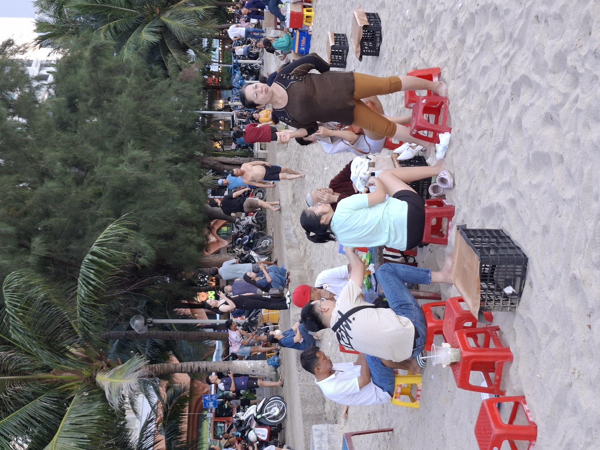 This media file captures a bustling scene on a sandy beach, likely in Bac Nha Trang Ward, Vietnam. The image is oriented vertically, showing a diverse group of people engaged in various activities. Several individuals are seated on low, red plastic stools arranged on the sand, some appearing to be socializing or enjoying drinks from clear plastic cups. Others are standing or walking, with a noticeable presence of motorbikes parked along what appears to be a walkway or road bordering the beach.
The background features lush green trees, including palm trees, suggesting a tropical or subtropical environment. In the distance, buildings can be seen, indicating a developed coastal area. The ground is covered in light-colored sand, with a few shells scattered around. The overall atmosphere seems lively and casual, with people dressed in everyday clothing, including t-shirts, shorts, and jeans. Some are barefoot, enjoying the beach setting.
One woman in the foreground, wearing a brown top and orange shorts, is looking towards the camera with a slight smile. Another person in a light blue shirt is also prominently featured. The presence of multiple people suggests a public gathering or a popular spot. The time of day appears to be daytime, possibly late afternoon or early evening, given the lighting and the relaxed activities. There is no clear indication of the weather, but the presence of many people outdoors suggests it is likely pleasant.
Visible text includes what appears to be "Tiger" on a blue box or cooler. The scene conveys a sense of community and leisure, with people making the most of a warm, beachside environment.