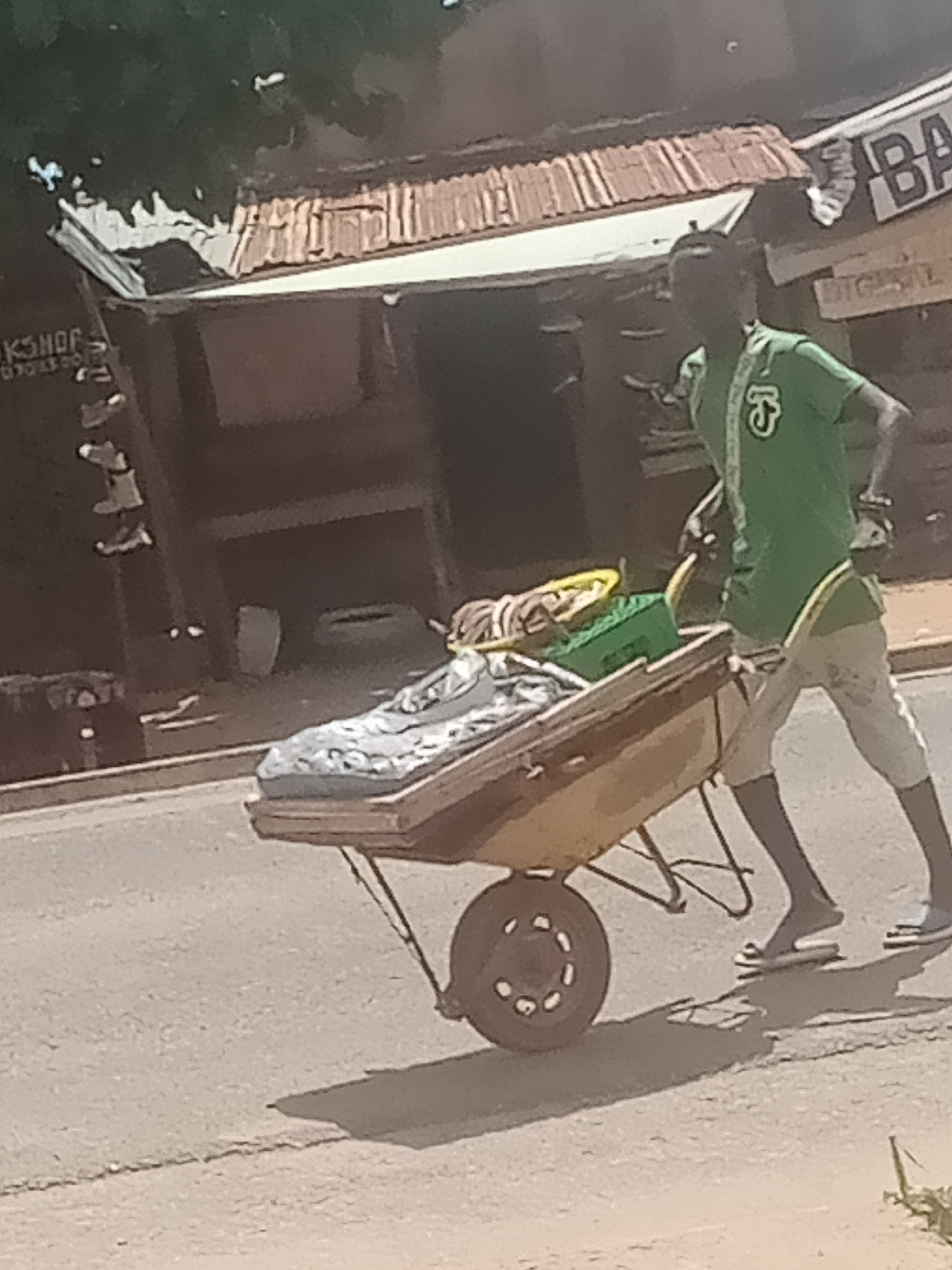 In Jalingo, Nigeria, a person wearing a green t-shirt and light-colored shorts is pushing a yellow wheelbarrow filled with various items, including what appears to be fabric and possibly food items covered by a grey patterned sheet. The wheelbarrow has a single wheel and is on a paved road. In the background, there are buildings with corrugated tin roofs, suggesting a market or commercial area. One building has signage that includes the letters "BA" and "BIGSAL". The overall scene depicts a street vendor or someone transporting goods in an urban environment.