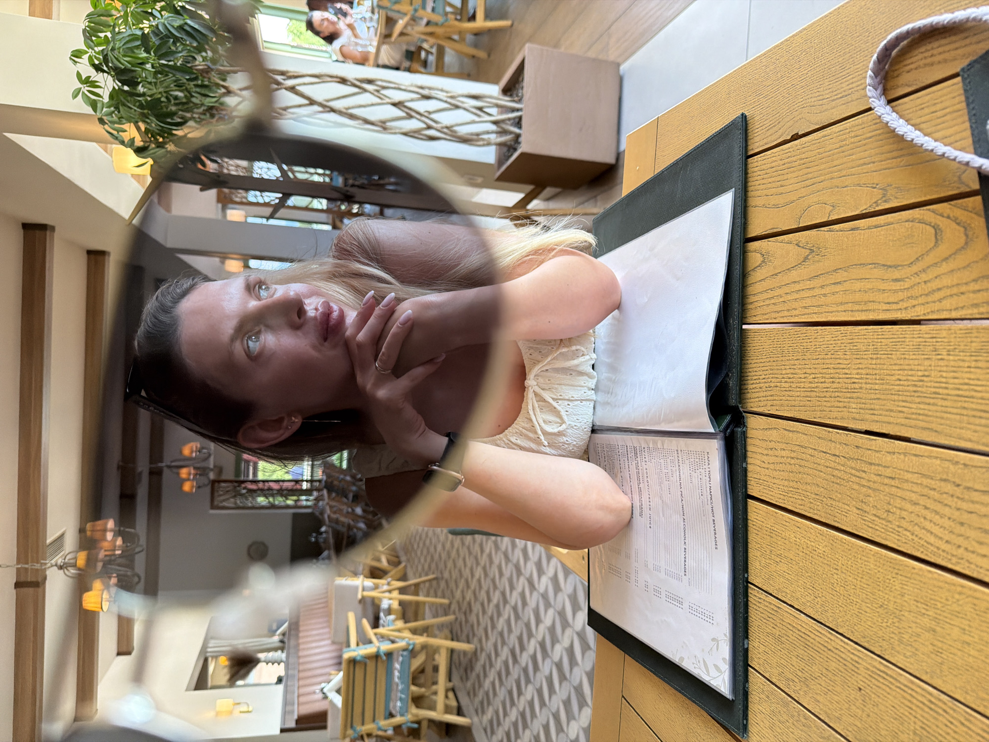 The image shows a woman sitting at a table in a restaurant, possibly in the city of Bigor, Montenegro. She is wearing a white dress and has her hands clasped in front of her. A menu is open on the table in front of her. In the background, other tables and chairs are visible, as well as a large potted plant. The restaurant appears to be modern and well-lit. The woman is looking off to the side, with a thoughtful expression on her face. The image is taken from a close-up perspective, focusing on the woman and the table.