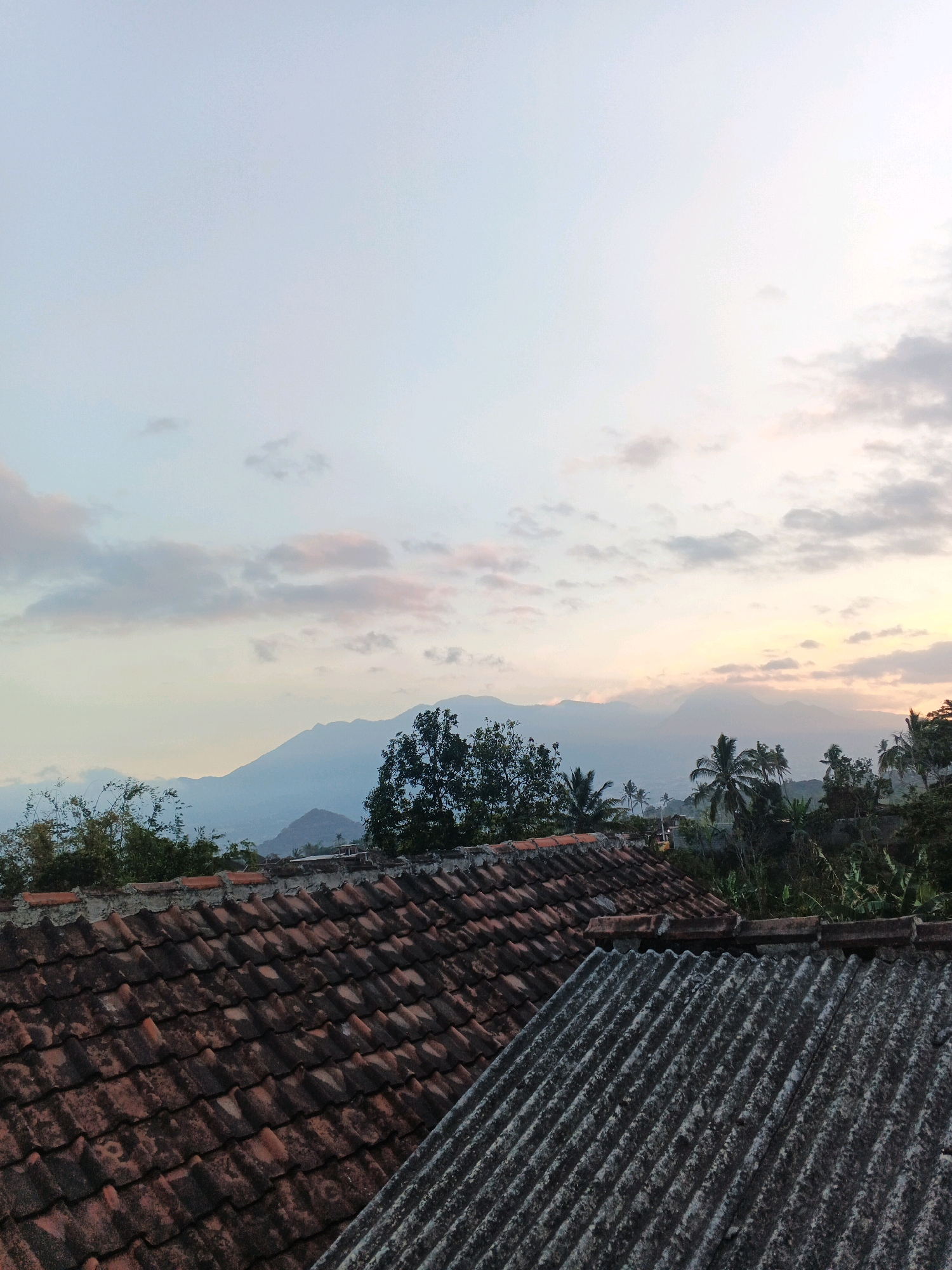 The image depicts a tranquil scene from Bocek, Indonesia, likely during the early morning or late evening. In the foreground, two rooftops are visible. The closest one is covered in weathered, reddish-brown clay tiles, showing signs of age and exposure to the elements. Adjacent to it is a roof made of corrugated material, appearing grayer and possibly made of asbestos or a similar substance.
Beyond the rooftops, a lush landscape unfolds. Dense green foliage and trees, including palm trees, are scattered across the terrain. In the middle ground, a series of rolling hills and mountains are silhouetted against the sky. The mountains appear hazy and distant, their peaks softened by the atmospheric conditions.
The sky is a prominent feature, displaying a gradient of soft colors. Near the horizon, a warm, yellowish-orange hue suggests the presence of the sun, either rising or setting. This light is diffused by scattered clouds, which range from light gray to soft pinks and blues, creating a serene and picturesque sky. The overall impression is one of a peaceful rural or semi-rural setting in Indonesia. There are no people or animals visible in the image.