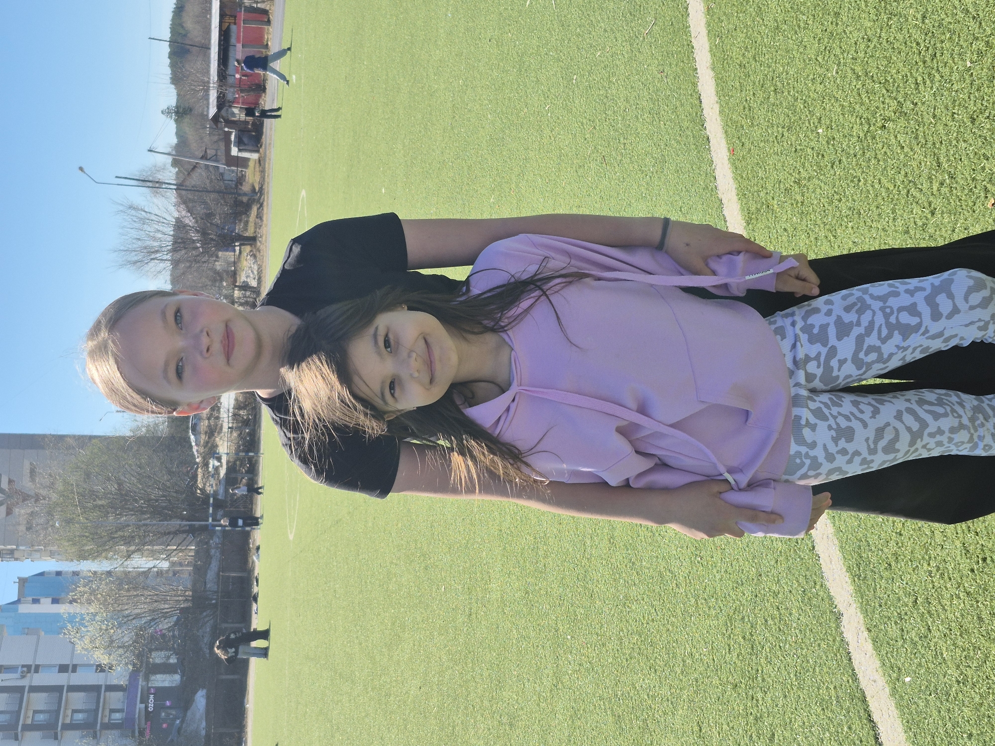Two young girls pose on a green field under a bright sky. One girl wears a black shirt and the other is in a light purple hoodie and patterned leggings. The location appears to be a recreational area with buildings and trees visible in the background, likely in a park or near a sports field in Chaikovsky, Russia. Other people are visible in the distance, further indicating a public space.
