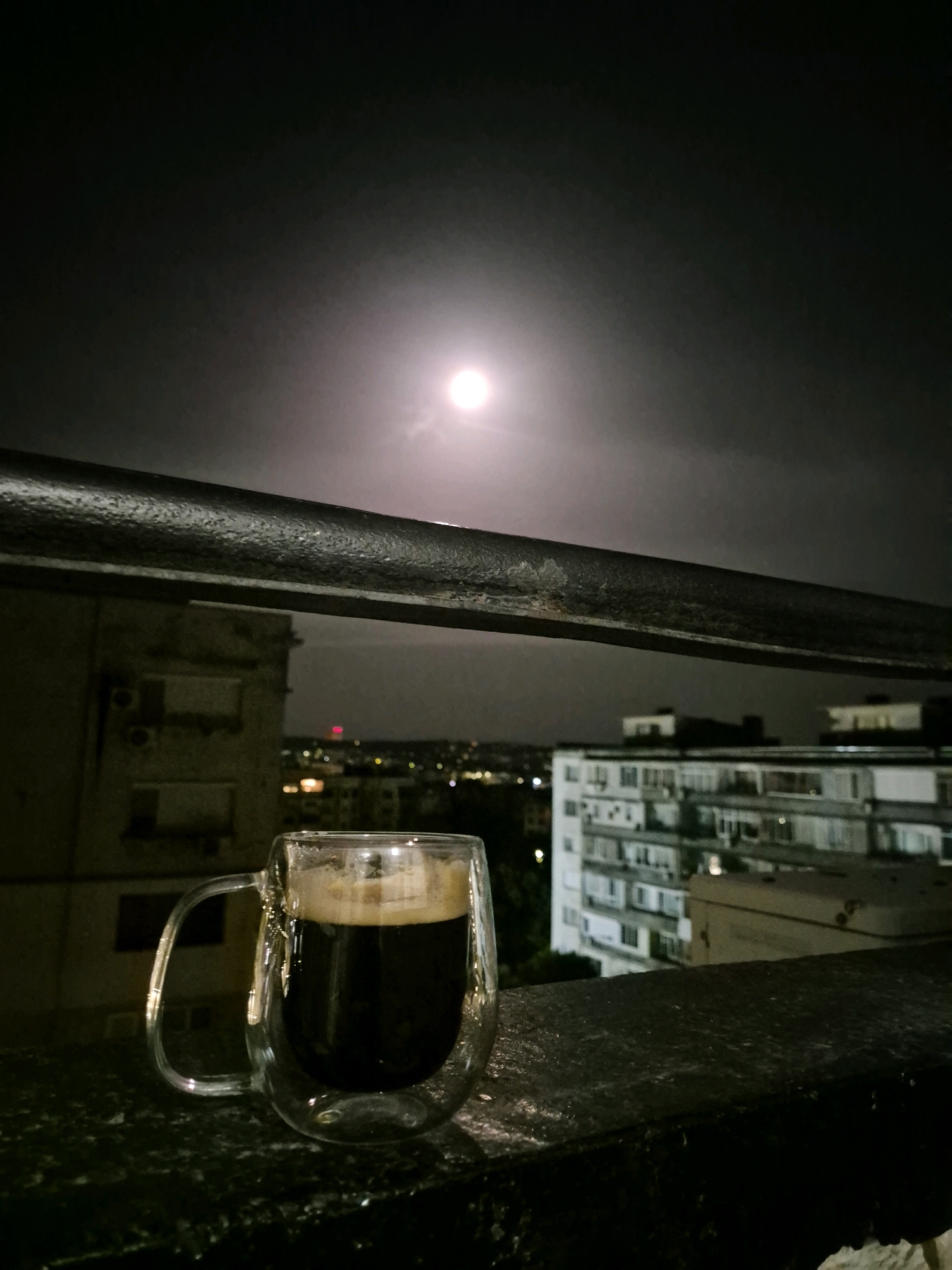 A clear glass mug filled with dark liquid and topped with a foamy layer sits on a dark, wet surface, likely a balcony railing. Behind the mug, a cityscape is visible at night, illuminated by distant lights. In the background, large, multi-story buildings with visible windows are prominent, with the sky above a dark, overcast gray. A bright, diffused light source, likely the moon through clouds, is visible in the upper part of the frame. A thick, dark metal railing runs horizontally across the middle of the image. The overall atmosphere is dimly lit and moody, suggesting a quiet evening scene.