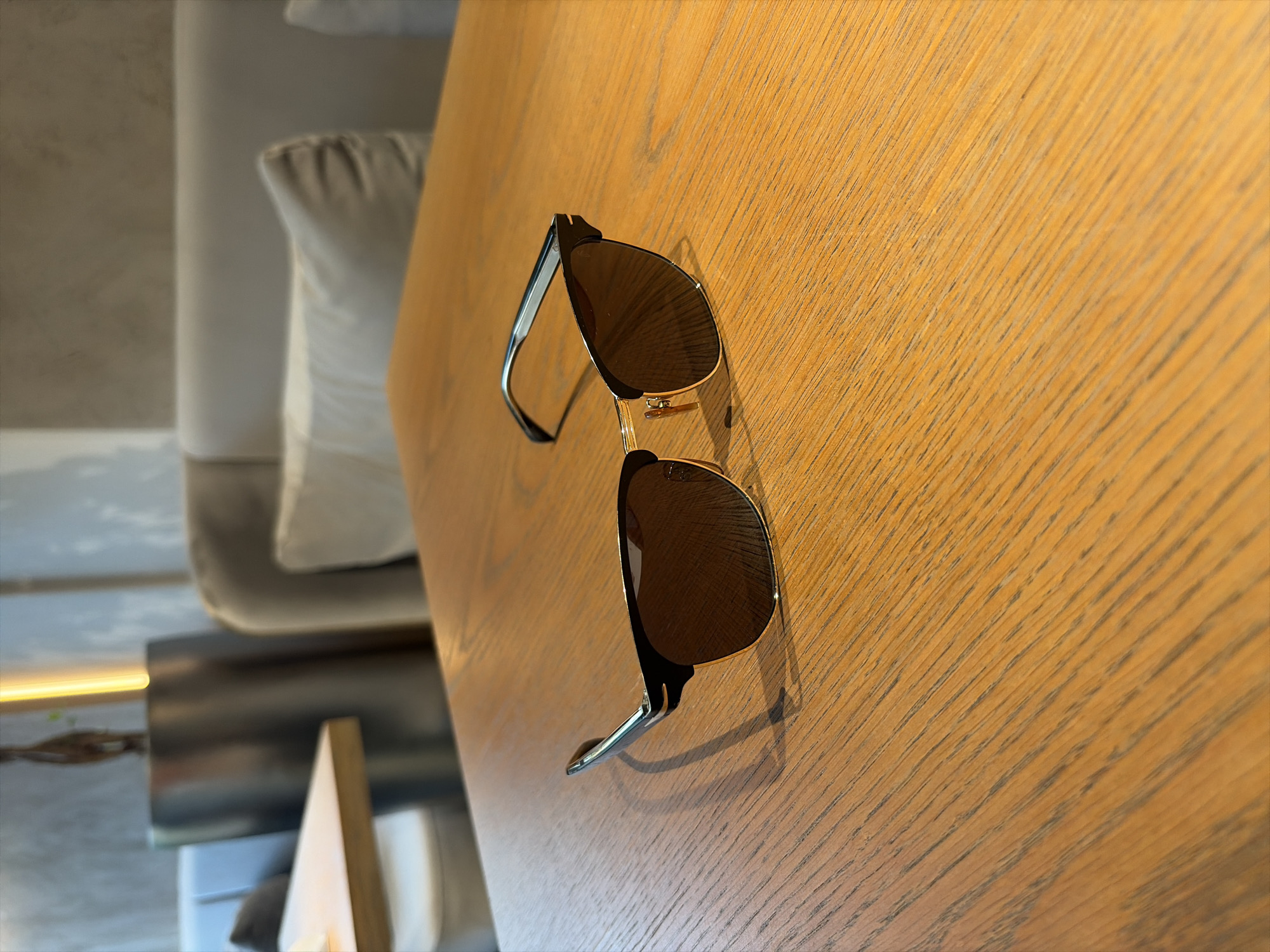 A pair of black and gold-rimmed sunglasses with brown lenses rests on a light brown wooden table. The sunglasses are positioned in the center of the frame, angled slightly towards the right. The table has a visible wood grain pattern. In the background, a blurred scene depicts what appears to be an indoor seating area, possibly a cafe or restaurant, with hints of upholstered furniture and a metallic cylindrical object. The lighting suggests an indoor environment. There is no visible indication of the location being Moscow, Russia.