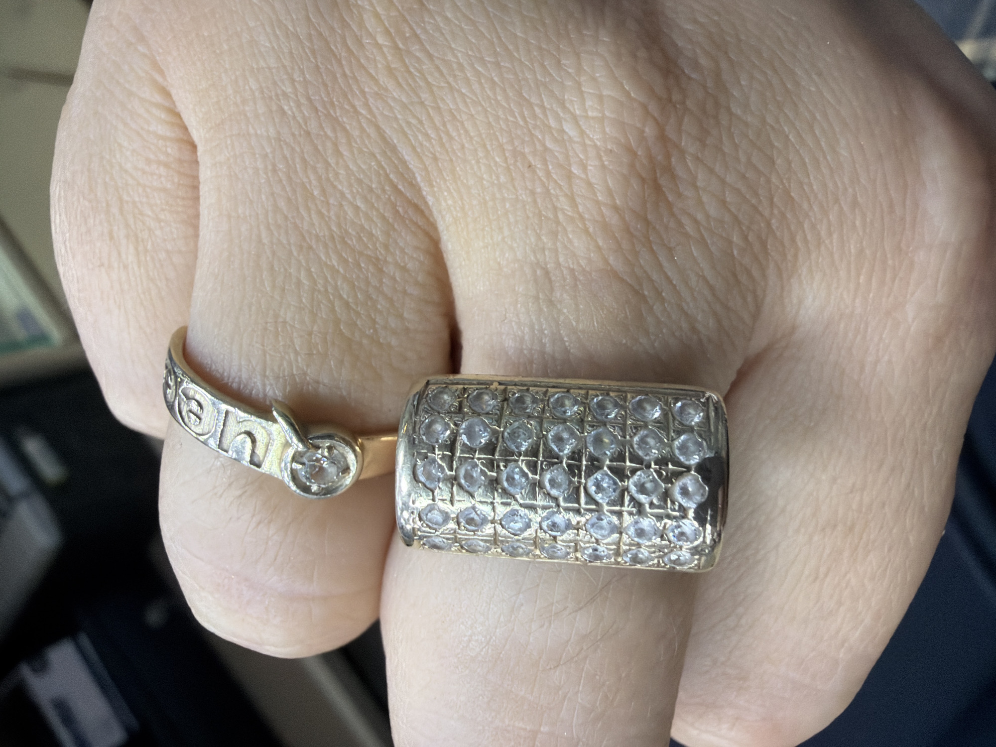 A close-up photograph captures a person's hand, specifically a finger, adorned with two distinct rings. The skin of the hand shows detailed texture, fine wrinkles, and some hair follicles, indicating an adult.
On the left side of the finger, a slender, golden-toned ring is visible. This ring features cut-out letters, with "en" or "eh" clearly discernible, suggesting part of a word or name. A small, round, clear gemstone, likely a diamond, is set into this ring.
The second, more prominent ring is rectangular or barrel-shaped, crafted from a similar golden-toned metal. Its entire surface is encrusted with numerous small, round, clear gemstones, possibly diamonds or cubic zirconia, arranged in a grid pattern, creating a sparkling effect.
The background is out of focus, appearing to be an indoor setting with indistinct dark and light surfaces, potentially a desk or table. The lighting is soft and even, highlighting the jewelry. No specific activity is depicted other than the display of the rings. No emotions are discernible from the hand alone. The provided location context is Sochi, Russia, but there are no visual cues within the image to confirm this.