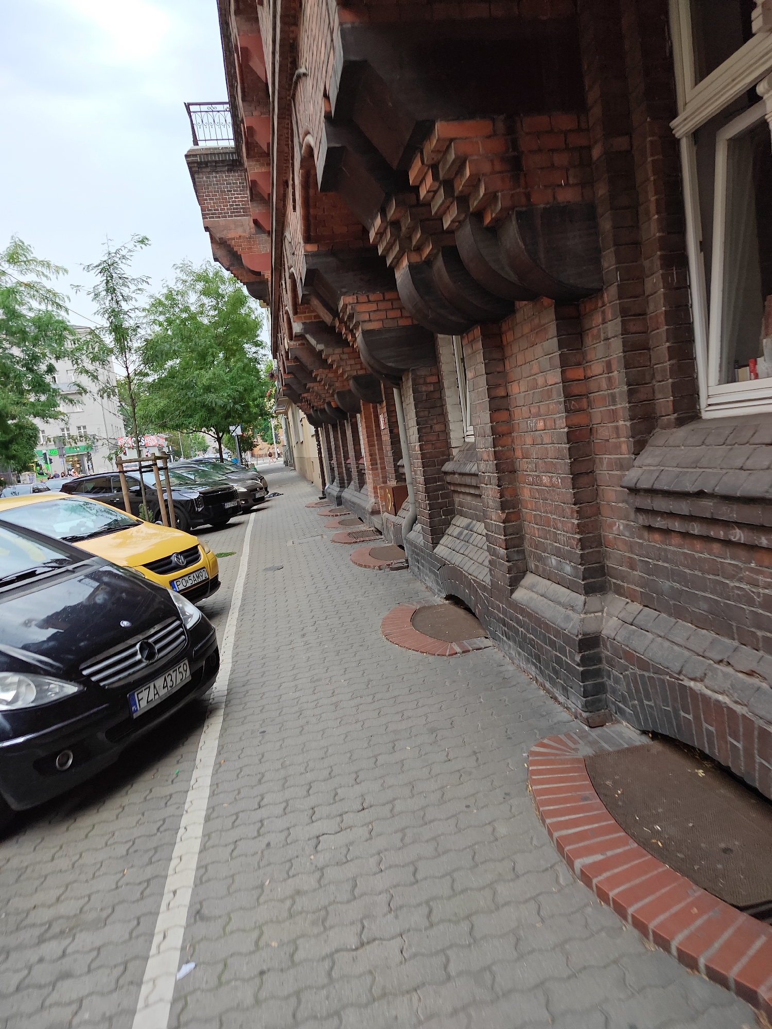 The image shows a street in Poznan, Poland, with cars parked along the sidewalk. The building on the right is made of brick and has a distinctive architectural style. The sidewalk is paved with interlocking concrete blocks. The weather appears to be overcast.