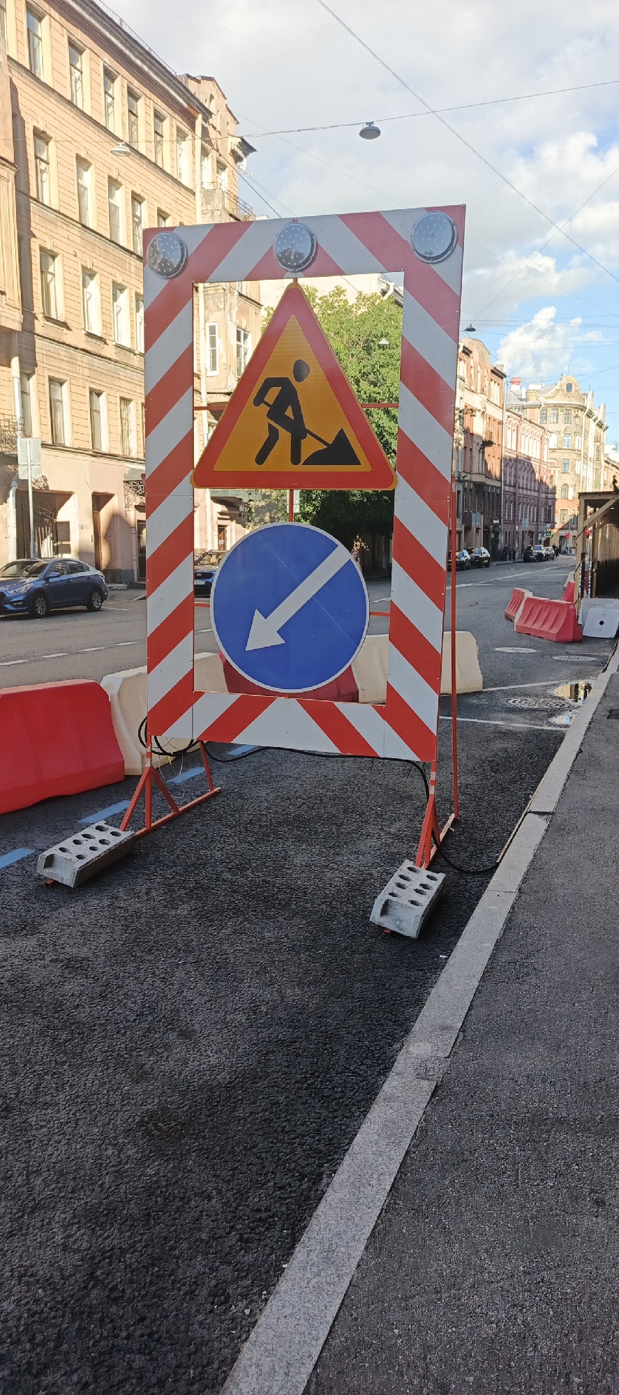 The image depicts a street scene in Saint Petersburg, Russia, likely during daytime, under a cloudy sky. In the foreground, a construction barrier is set up, featuring a prominent red and white striped frame. Mounted on this frame are two traffic signs: a yellow triangular sign with a black silhouette of a person digging with a shovel, indicating road work, and a blue circular sign with a white arrow pointing diagonally downwards and to the left, signifying a mandatory direction of travel.
The barrier is supported by orange metal legs resting on a newly paved asphalt surface. Gray concrete blocks with circular indentations serve as weights for the barrier. Red plastic barriers are placed on either side of the construction area, delineating the work zone.
In the background, a blue car is visible on the street. Beyond the road, historic multi-story buildings with arched windows and decorative facades line the street, typical of Saint Petersburg's architecture. Overhead, utility wires and streetlights are present. The overall scene suggests ongoing road maintenance or construction in a developed urban environment.