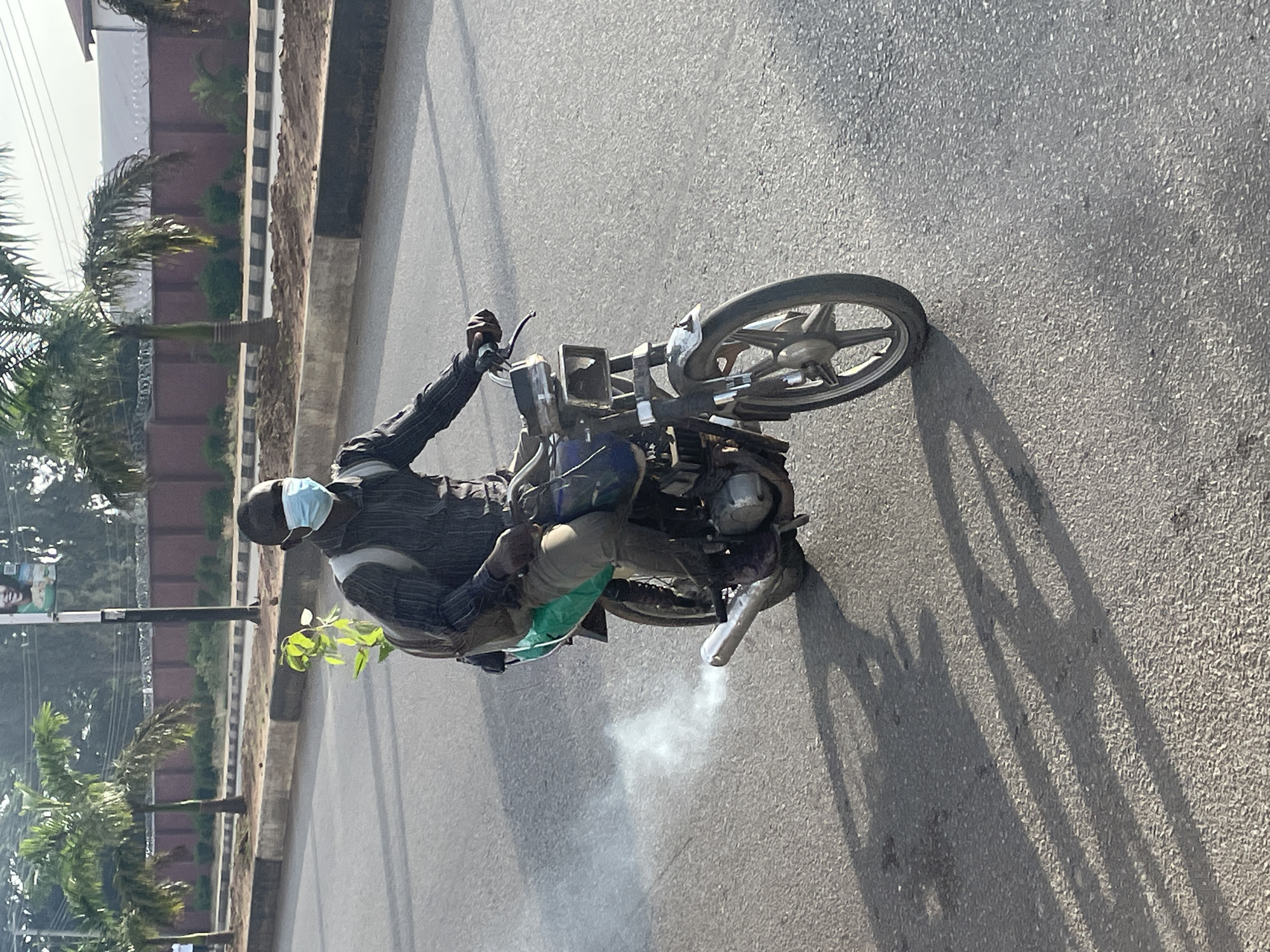 A person wearing a light blue face mask and a dark striped long-sleeved shirt is riding a motorcycle. They have a light-colored backpack on, and a green bag is strapped to the back of the motorcycle. A small plant with green leaves is attached to the backpack, extending upwards. The motorcycle is emitting a visible plume of white smoke from its exhaust pipe, which is pointing downwards and to the left. The motorcycle has a metal fender on the front wheel and a silver exhaust pipe. The scene appears to be on an asphalt road with visible tire tracks and shadows cast by the motorcycle and rider. In the background, there are palm trees and a dark red wall with barbed wire on top. A street sign with a picture of a smiling person is visible above the trees. The environment suggests an outdoor setting, likely a street in an urban area, possibly Jos, Nigeria.