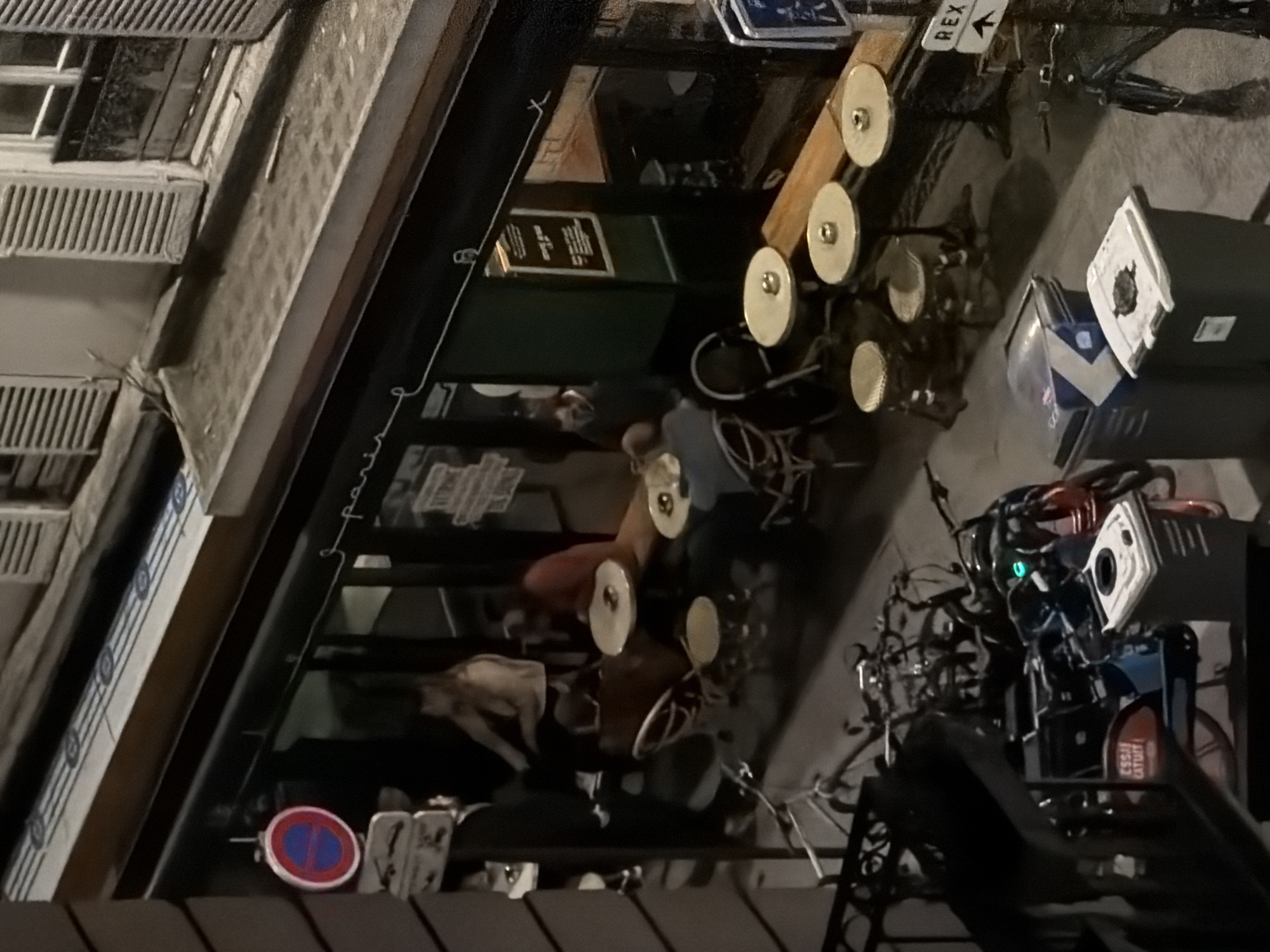 Here is a detailed description of the image:
The image shows a street scene in Paris, France, likely at night, taken from an elevated position. Visible are the exteriors of buildings with traditional Parisian architectural details, including windows with shutters. A street-level establishment, possibly a cafe or restaurant, is present with tables and chairs set up on the sidewalk. People are seated at some of the tables, and there appears to be interaction among them. A "no parking" sign is visible, along with miscellaneous items on the sidewalk. Bicycles are parked nearby. The overall lighting suggests it is either dusk or night, with artificial lighting illuminating the scene.