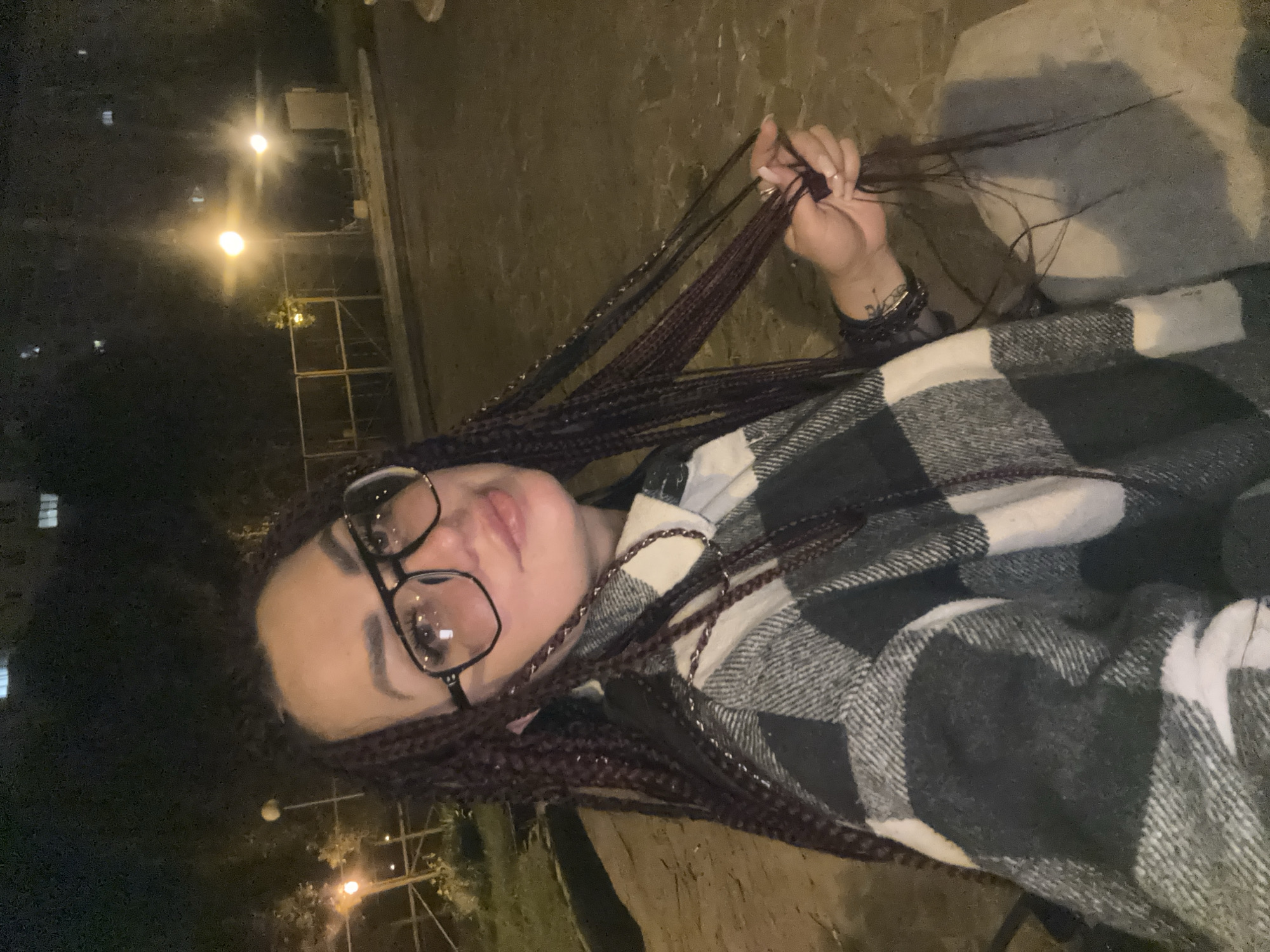 A young woman with dark braided hair and glasses is captured in a close-up, waist-up shot. She is wearing a black and white plaid shirt. Her right hand is raised, holding a section of her braids, and she has several bracelets on her wrist. The lighting suggests it is nighttime, with artificial lights visible in the background, possibly streetlights or lights from a building. The ground appears to be paved or made of concrete. There are some shadowy areas and a general sense of an outdoor urban environment at night. The woman's expression is neutral or slightly posed for the photo.