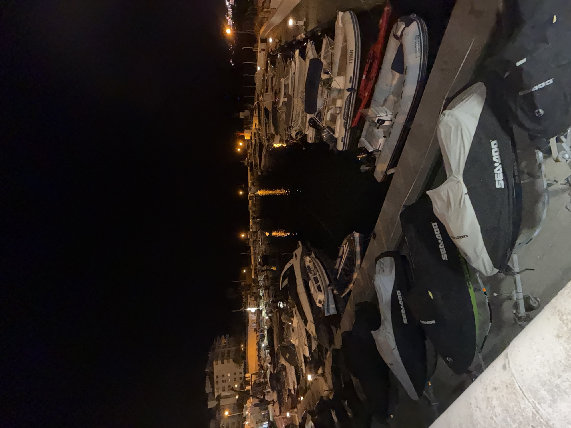 The image shows a nighttime view of a marina in Palmanova, Spain. The scene is illuminated by artificial lights, creating reflections on the dark water. Several boats are docked in the marina, with some covered and others uncovered. In the foreground, a row of jet skis are parked on a paved surface, many of them covered. Some of these jet skis have the brand name "SEA-DOO" visible on their covers. In the background, buildings and streetlights are visible, indicating the presence of a town or city. The overall atmosphere is calm and serene, typical of a harbor at night.