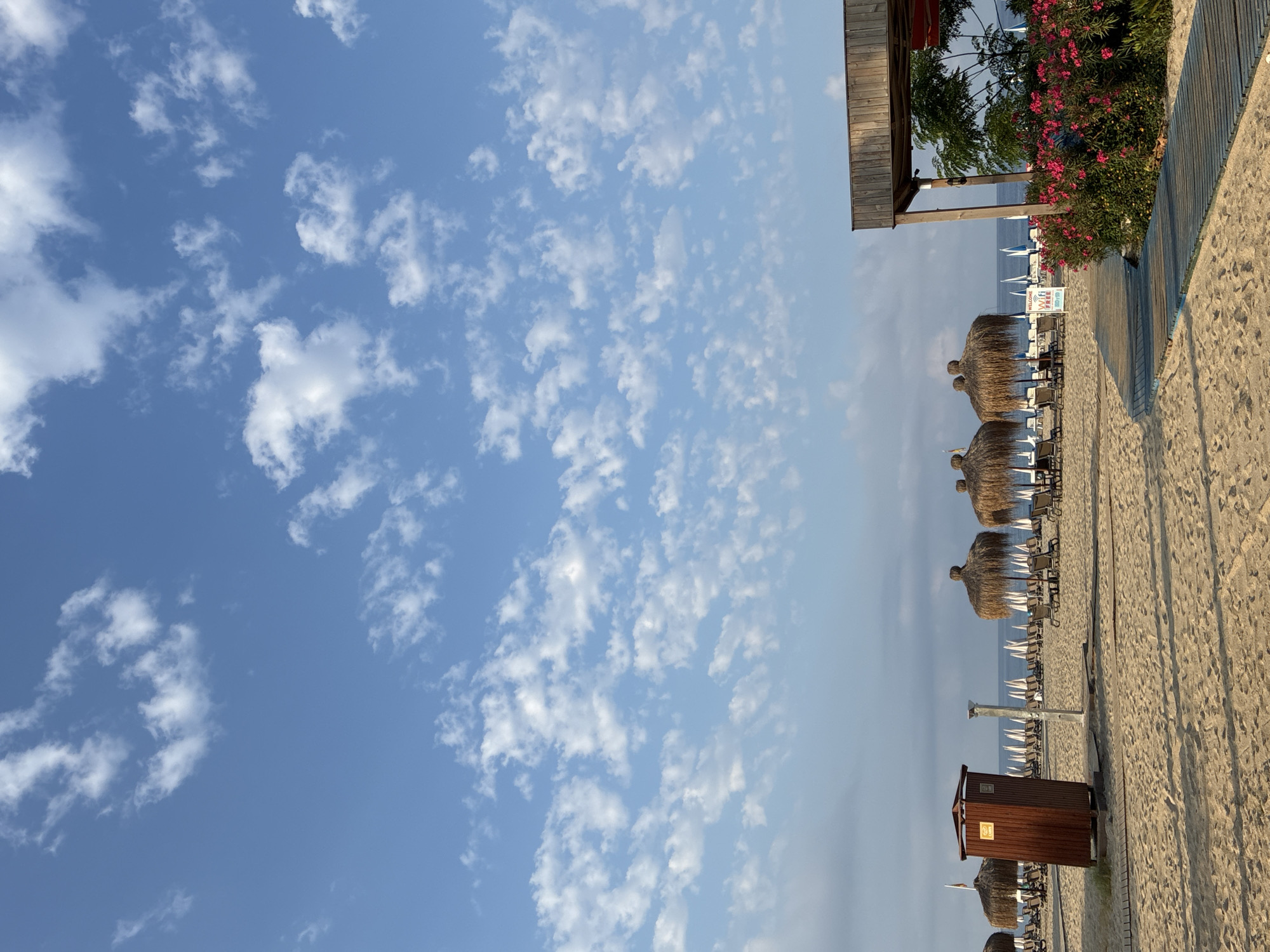 The image depicts a beach scene in Alanya, Turkey, under a bright blue sky dotted with scattered white clouds. The foreground is dominated by a sandy beach with a wooden boardwalk curving to the right. To the left, a dark wooden changing booth or restroom stands on the sand, accompanied by a shower fixture. Further back, rows of thatched umbrellas and lounge chairs are set up on the beach, with the calm sea visible in the distance. On the right side of the boardwalk, a wooden structure shades a vibrant bush with pink flowers. The overall atmosphere suggests a resort or public beach area prepared for visitors.