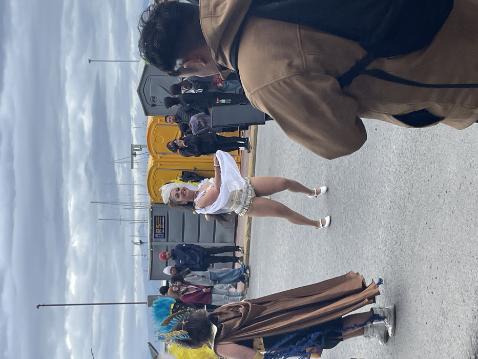 The image captures an outdoor event, possibly a parade or festival, under an overcast sky. The setting appears to be a waterfront area, with numerous boat masts visible in the background against the grey, cloudy sky.
In the immediate foreground, a person is seen from the back and side, wearing a brown hooded jacket or coat and a black backpack. Their head is turned slightly to the left, observing the scene.
The central focus of the image is a woman dressed in a vibrant performance costume, striking a pose or mid-dance. She is smiling broadly and looking directly towards the camera. Her attire includes a white, off-the-shoulder top with flowing fabric, a decorative short bottom garment in white and gold hues, and white ballet-style flat shoes. She has long dark hair and wears a white headpiece. Her legs are flexed, indicating movement.
To the left of this woman, partially obscured and viewed from behind, is another individual in an elaborate costume. This person is wearing a large, colorful feathered headdress, primarily featuring blue, yellow, and brown feathers, and a long, flowing brown cloak-like garment. They are wearing metallic silver shoes.
In the midground, behind the performing woman, a line of bright yellow portable toilets is visible. In front of and around these facilities, several other individuals are present, dressed in everyday clothing, appearing to be spectators or participants. Some are looking towards the performers, and at least one person in the group is smiling.
On a grey utility box or structure located near the portable toilets, the letters "RS" are visible. The ground beneath the people is paved, resembling asphalt or concrete. The lighting indicates it is daytime, despite the cloudy weather.