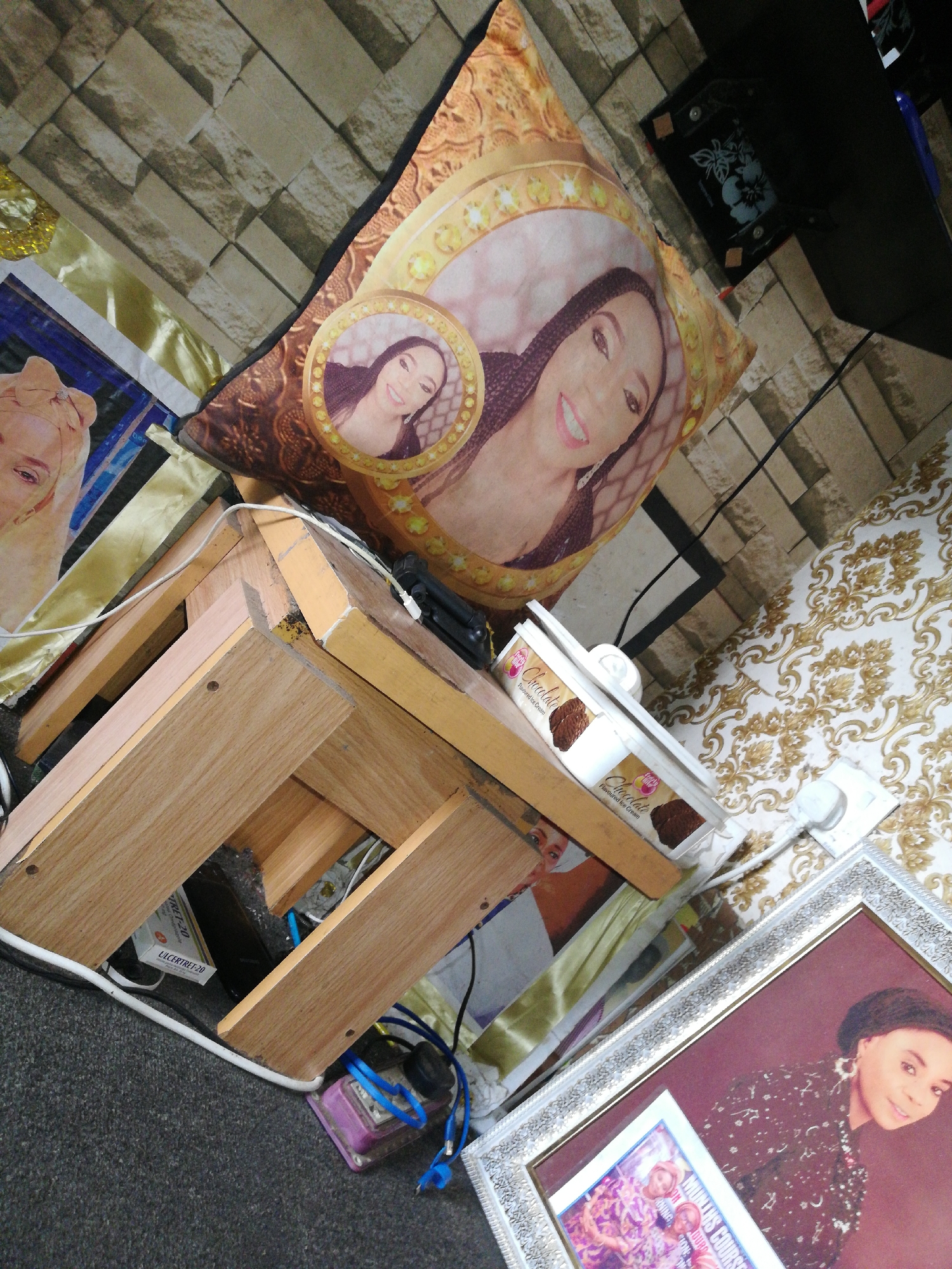 A cluttered interior scene in Lagos, Nigeria. The room features a light brick-patterned wall and patterned wallpaper in beige and gold. A small, wooden stool with a light-colored surface supports various items. Resting on the stool is a pillow with a portrait of a woman, and a plastic container labeled "Chocolate Flavoured Ice Cream." Scattered beneath the stool are electrical cords, a purple electronic device, and a small package with the text "ULCERTRET-20." The floor is covered with dark carpet. A framed portrait of a woman and another framed picture are positioned towards the bottom right of the frame. There are also other random materials present in the space.
