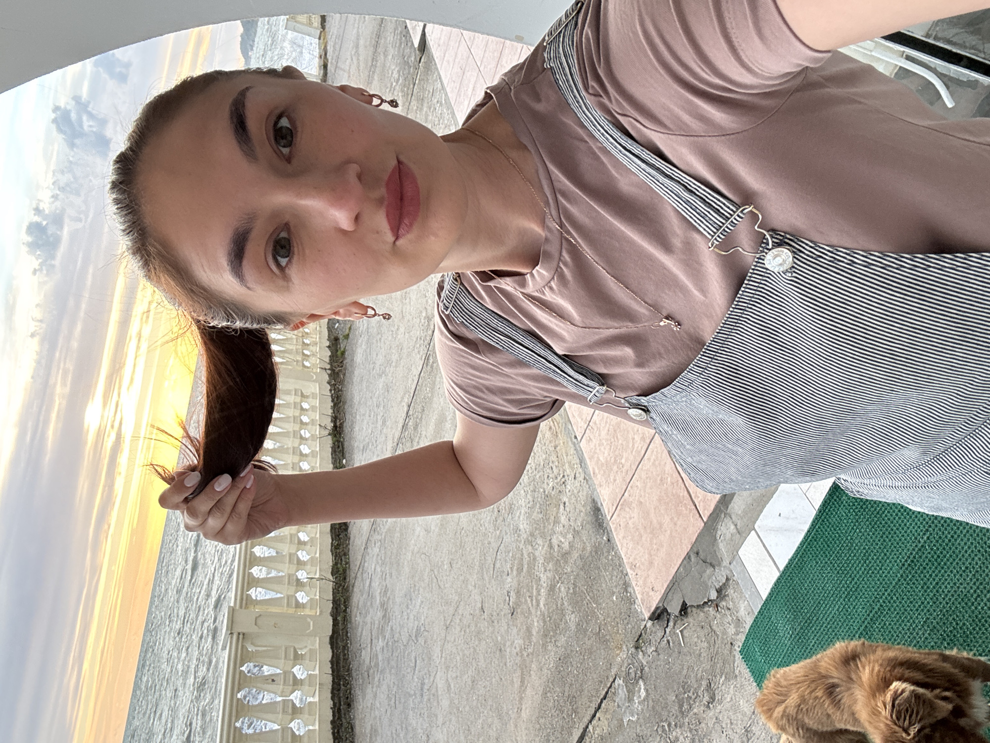 A woman stands outdoors, holding a lock of her brown hair in her left hand. In the background, the sea is visible with a sky indicating either sunrise or sunset. The location appears to be a waterfront area, possibly in Sochi, Russia, based on the architectural style of the railing and the general environment. The woman wears a light brown t-shirt and striped overalls. A small part of a brown dog is seen on the lower-right corner. The ground is paved with concrete. The photograph is taken at an angle, making the scene appear tilted.