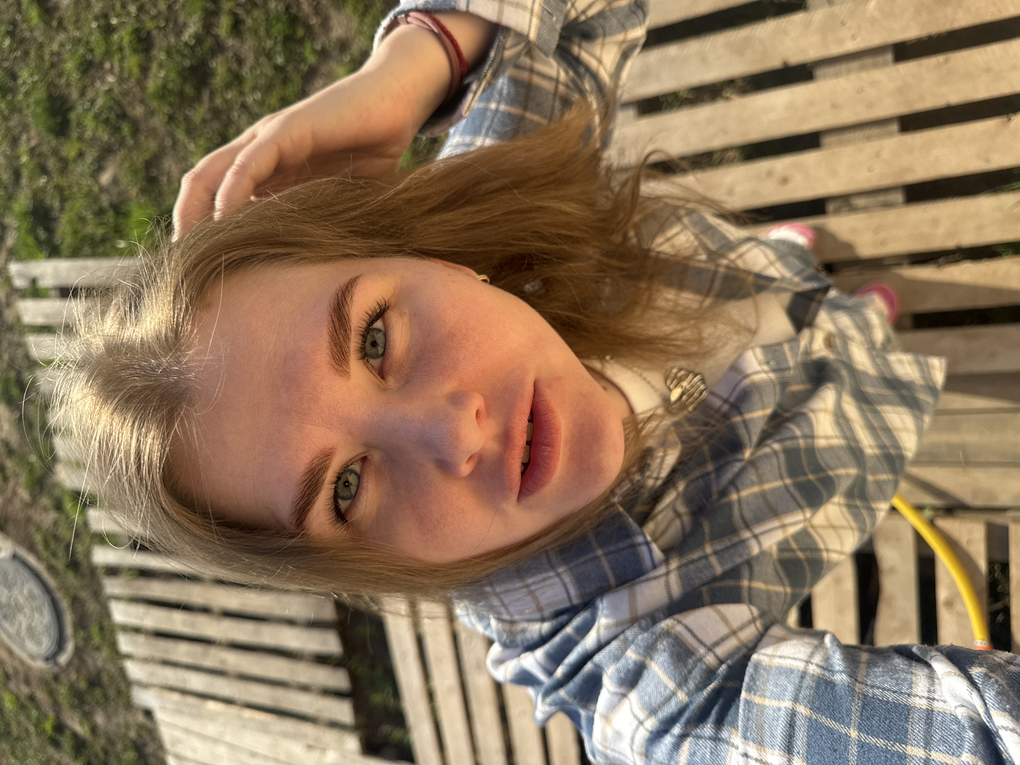 A young woman is pictured in a selfie, seemingly outdoors on a wooden structure that appears to be a bench or a slatted surface. She is wearing a blue and white plaid shirt over a white top. Her blonde hair is partially obscuring her face on one side, with her hand touching her hair. The background shows some greenery, possibly grass or foliage. The scene is bathed in natural light, indicating it is daytime. The precise location within Ферма, Russia, cannot be determined from this image alone.