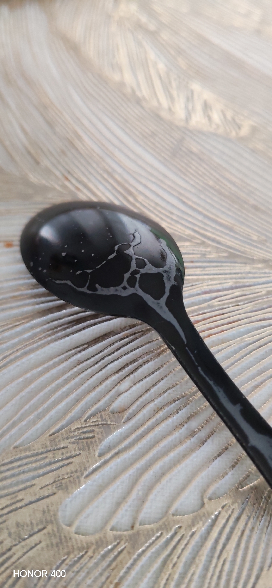 A close-up shot shows a black spoon resting on a surface with a textured, feather-like pattern. The spoon is slightly wet with visible water droplets and a network of water marks on its surface. The background texture consists of alternating raised and recessed lines, resembling feather contours, in a light cream color. The spoon is positioned diagonally across the frame, with the bowl of the spoon at the upper-left and the handle extending towards the lower-right. The bottom-left corner of the image contains the text "HONOR 400". There are no people visible in the image. The photo does not depict any information related to the city of Noviy, Russia.