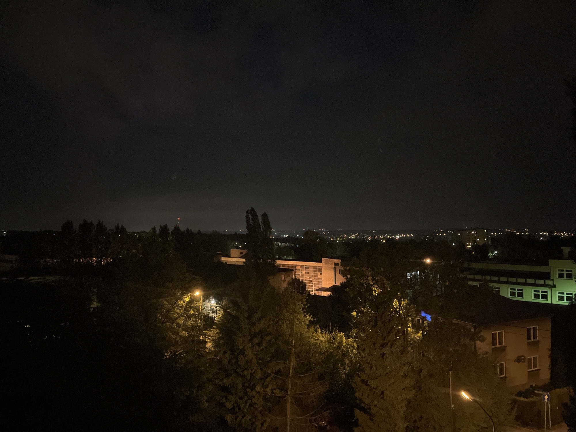 A nighttime, low-angle view from Unknown, Unknown. Visible are trees, buildings illuminated by artificial lights, and distant city lights under a cloudy sky. Streetlights cast a glow on the surrounding trees and buildings. The overall tone is dark, with pockets of illumination highlighting the urban landscape.