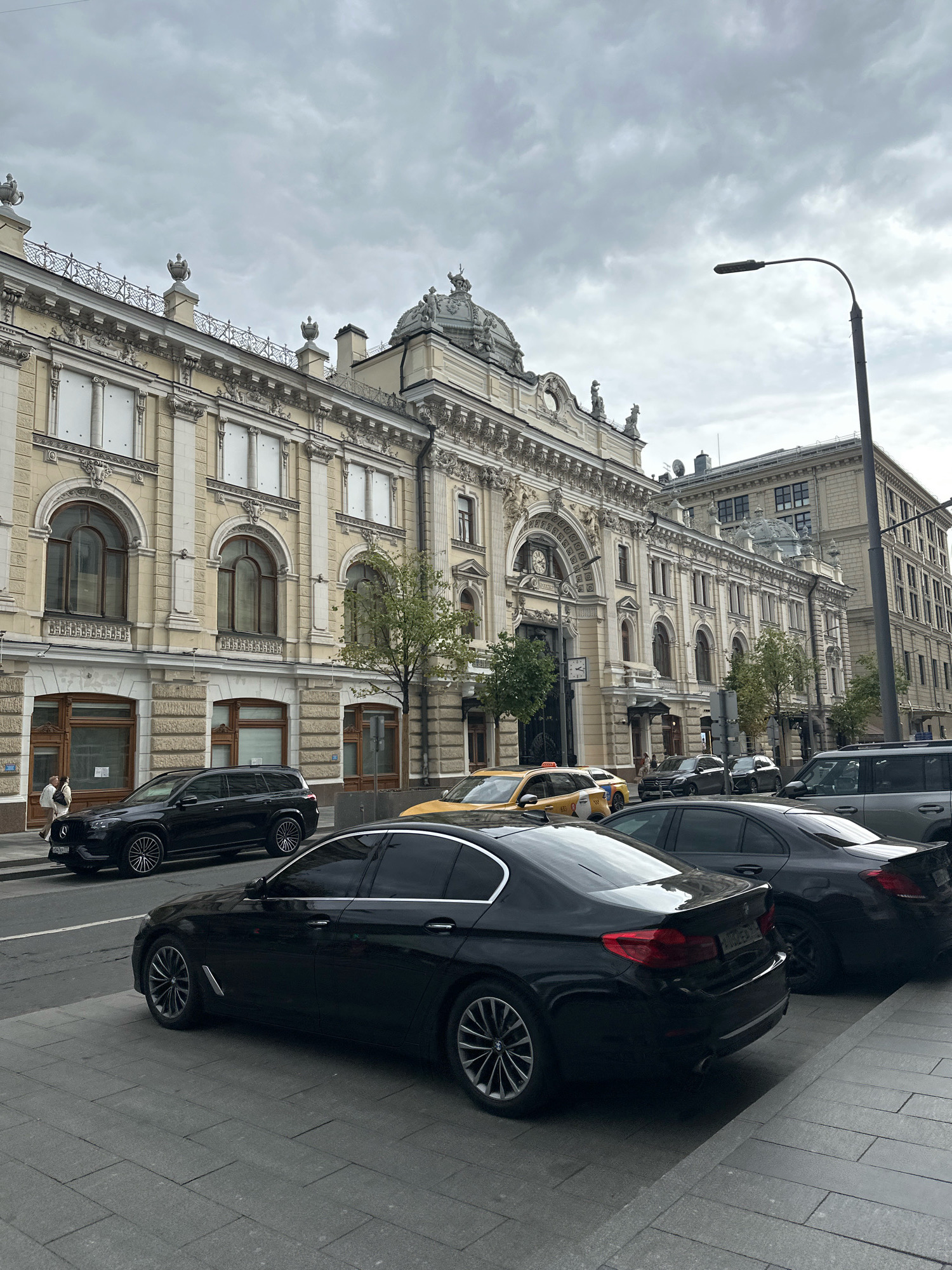 The image depicts a street scene in Moscow, Russia, with a prominent classical-style building lining the street. Several vehicles are parked and driving along the road, including black sedans, a black SUV, and a yellow taxi. The building features ornate architectural details, including arched windows, decorative carvings, and statues. The sky is overcast, suggesting a cloudy day. A street lamp is visible on the right side of the frame. Pedestrians are present on the sidewalk, with two individuals visible in the foreground near the black SUV. The street is paved with interlocking gray bricks. The overall impression is one of an urban environment with historic architecture and contemporary city life.