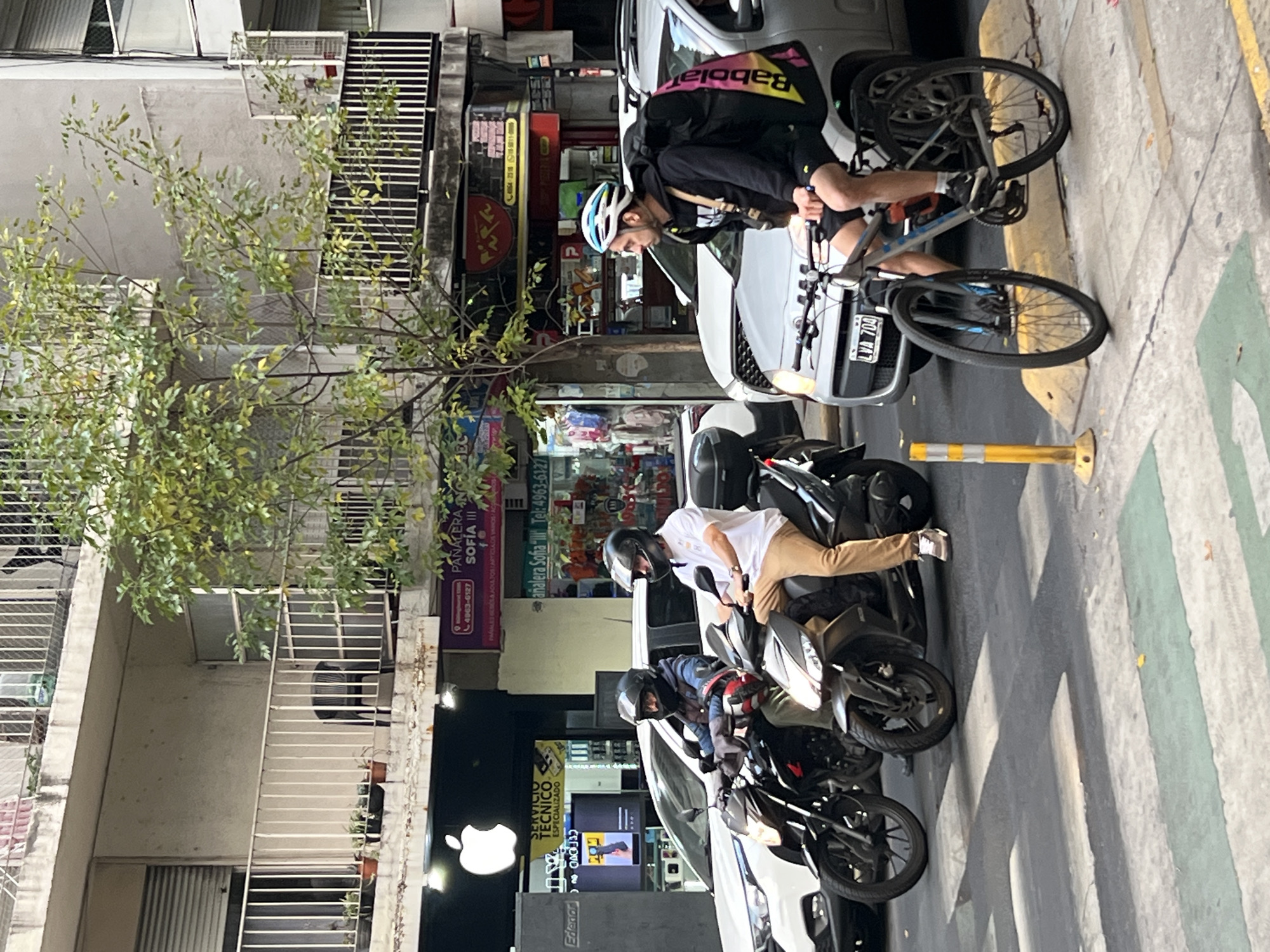 The image captures a street scene in Buenos Aires, Argentina. A cyclist wearing a helmet and a Babolat-branded backpack is standing with his bike near a parked white car. Further down the street, two people are seated on motorcycles. The motorcycles are parked in a row, partially obstructing the sidewalk. Adjacent to the parked motorcycles, an "Apple" logo is visible above a storefront featuring the signage "SERVICIO TECNICO ESPECIALIZADO" and "Edenar CA". The store also has a screen with a hand holding a computer part. Across the street, additional storefronts are visible, including one that says "PANALERA NOS SOFIA III". Signage also includes phone numbers.
