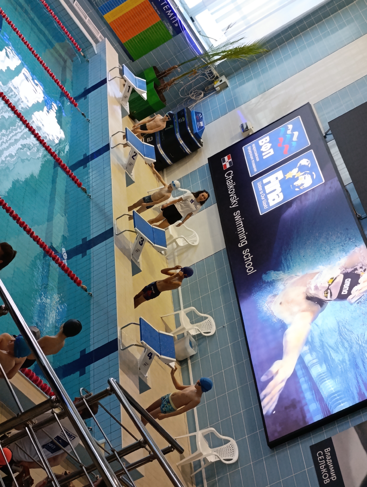 The image shows an indoor swimming pool, likely located in Chaikovsky, Russia. Several swimmers are standing on starting blocks numbered 1 to 4, preparing to dive into the pool, which is divided into lanes by red lane markers. A woman is standing near the swimmers, possibly a coach or referee. In the background is a large digital display showing promotional images for the "Chaikovsky swimming school", along with logos for the "All-Russian Swimming Federation" ("Воп") and "Fina", as well as an image of swimmer, Vladimir Selkov. A palm tree is visible near the display, and white plastic chairs are positioned near the starting blocks.