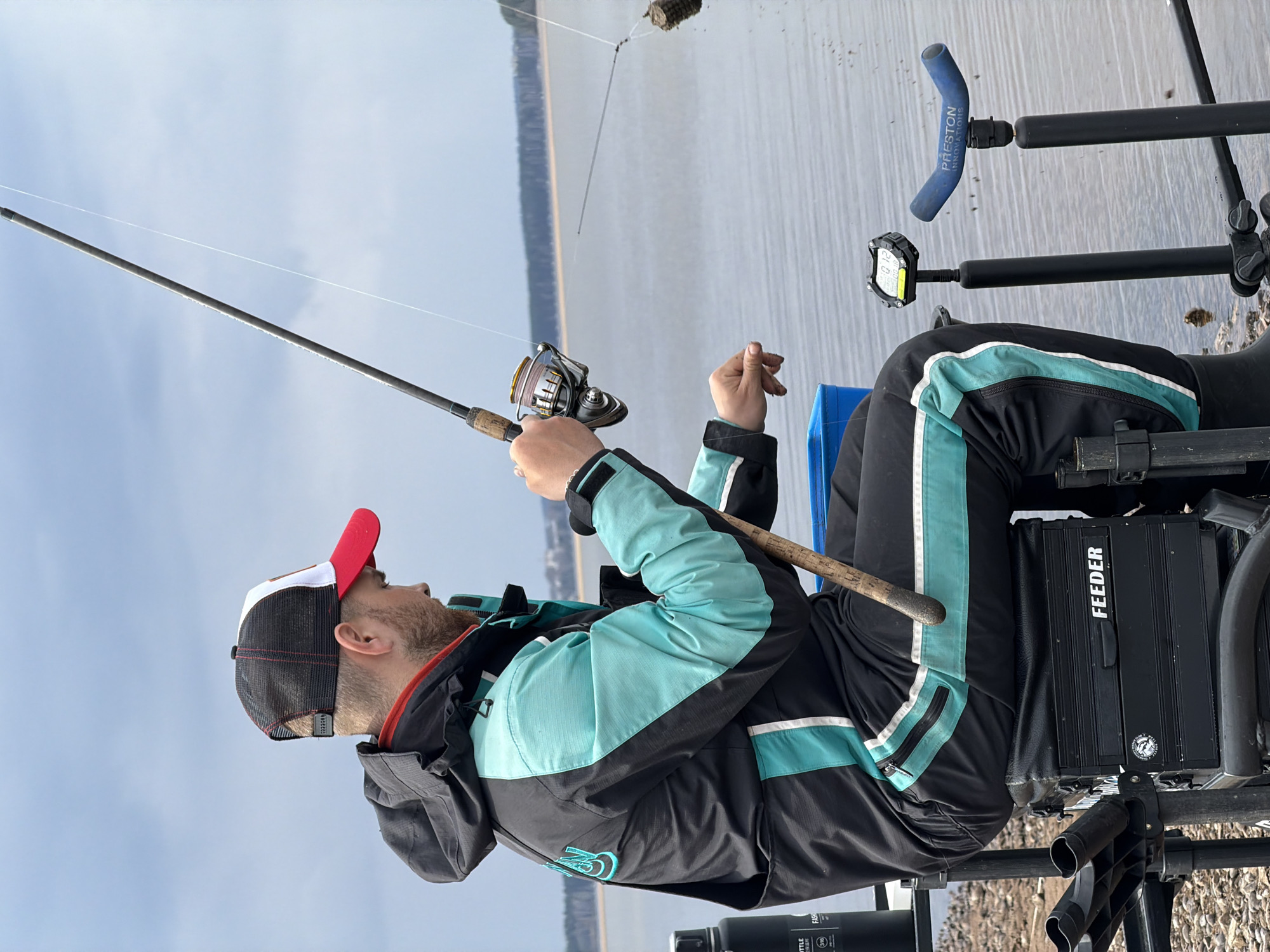 A man is fishing in a body of water in what is most likely Chaikovsky, Russia, evidenced by the brand "FEEDER" visible on his fishing box, a brand commonly associated with fishing in that region. He is seated on a fishing chair, holding a fishing rod with a reel. The line is cast into the water, with some kind of bait or weight attached to the end. The man is wearing a black and teal jacket, and a red, white, and black baseball cap. The sky is overcast. The shoreline is visible with various fishing equipment placed nearby.