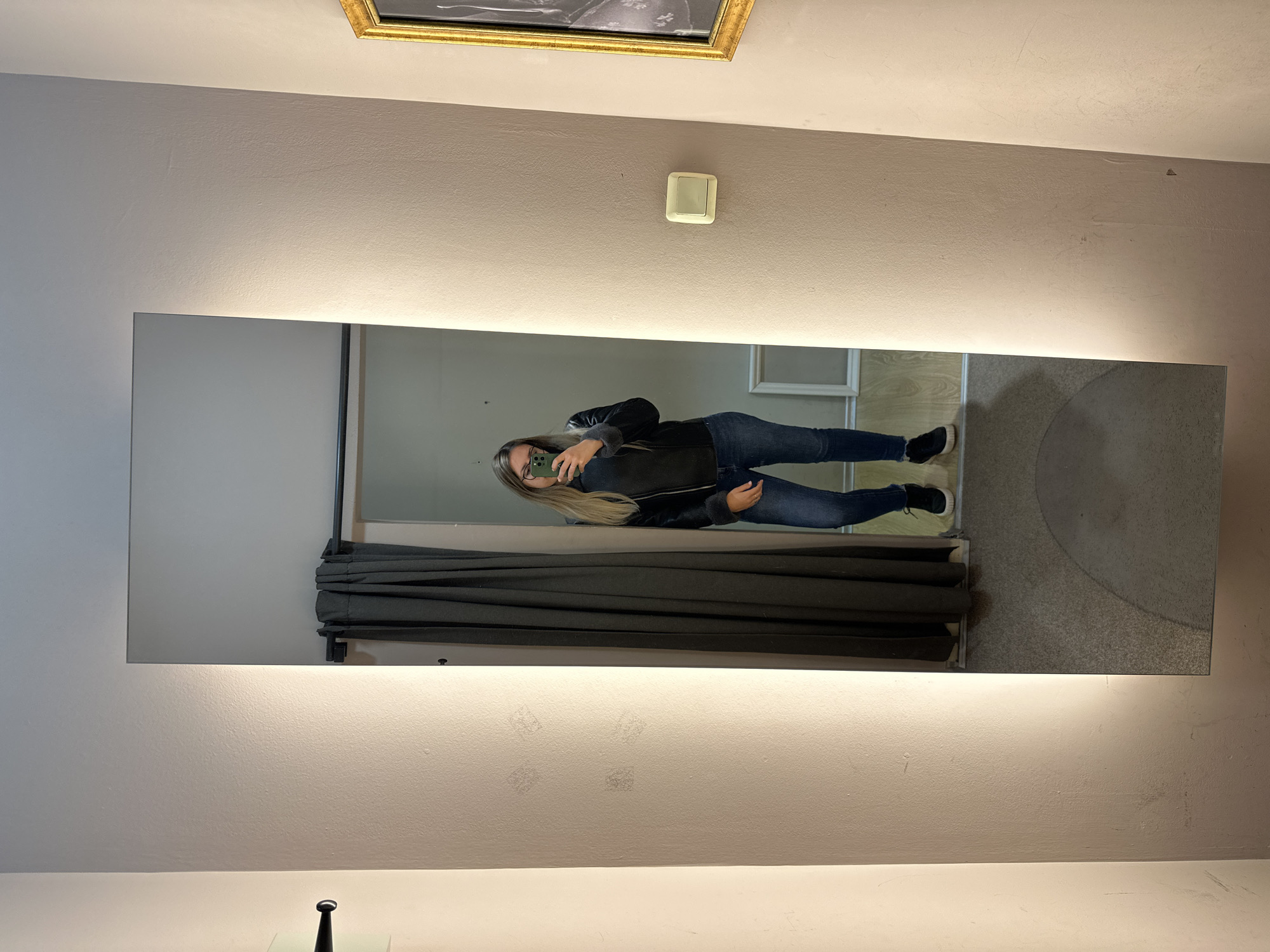 The image shows a vertical mirror with integrated lighting, mounted on a beige wall. The mirror reflects a woman taking a selfie with her phone. She is wearing a black jacket and blue jeans. Behind her in the reflection is a dressing room with dark gray curtains. Above the mirror, there is a light switch and a framed picture. The location is most likely a clothing store or similar retail environment. I cannot determine the specific location in Sofia, Bulgaria without additional context.
