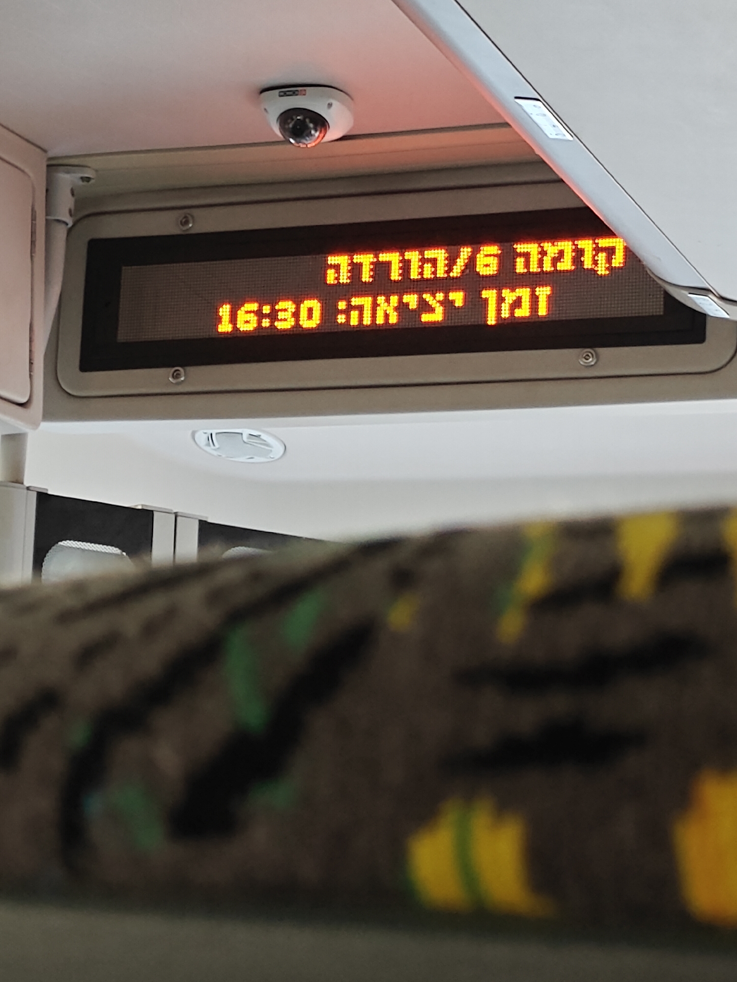The image shows an electronic display inside a vehicle, likely a bus or train, in Netanya, Israel. The display shows text in Hebrew and the time. The text reads "קדימה 6/הורדה" which translates to "Kadima 6/Drop-off" and below it "זמן יציאה: 16:30" which translates to "Departure Time: 16:30". The time displayed is 16:30. A small security camera is visible mounted on the ceiling above the display. The interior of the vehicle, including seats with a patterned upholstery, is partially visible in the foreground and background. The overall scene appears to be a functional interior of public transportation.