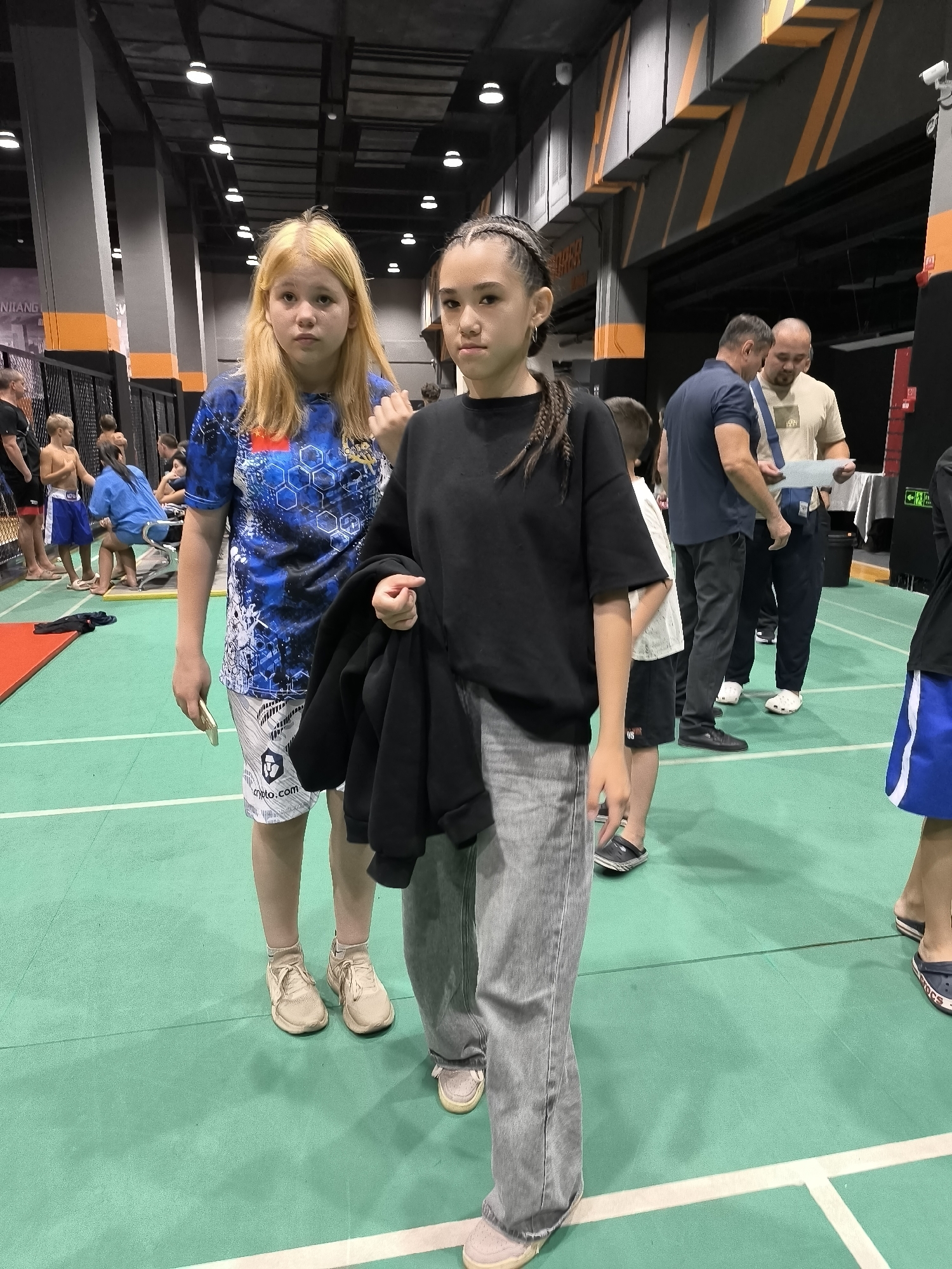 Two young females stand facing the camera on a green mat. The female on the left has blonde hair and is wearing a blue and white patterned t-shirt and white shorts. The female on the right has dark hair styled in braids and is wearing a black t-shirt and grey pants, with a black jacket draped over her arm. Behind them, several other people are visible, some standing and some sitting. The environment appears to be an indoor sports facility with a high ceiling and industrial lighting. There are also some black cages or enclosures visible in the background.