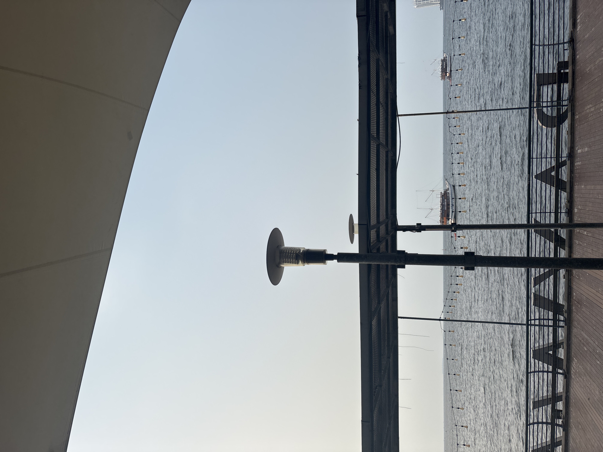 This image captures a scene at Ancol Beach City, North Jakarta, Indonesia. In the foreground, a wooden boardwalk runs horizontally across the bottom of the frame. Above the boardwalk, a metal railing with triangular supports is visible. Beyond the railing, the dark blue water of the ocean stretches out, dotted with small waves. A string of bare light bulbs is strung across the water, adding a decorative element.
In the mid-ground, two traditional wooden boats with multiple sails are visible on the water, sailing in the same direction. The sky above is a pale, hazy blue, suggesting it is daytime, possibly early morning or late afternoon given the soft light.
A dark metal structure, likely a canopy or part of a building, extends from the left and top edges of the image, casting a shadow and framing the view of the sea. Two tall lampposts with wide, conical shades stand prominently in the middle ground, positioned in front of the metal structure. The lampposts are dark grey and cylindrical.
In the far distance, on the right side of the image, a tall building or structure with a white sail-like design can be seen, indicating the presence of other buildings or landmarks in the vicinity. The overall impression is one of a waterfront promenade, designed for leisure and relaxation, with elements of both natural beauty and man-made structures.