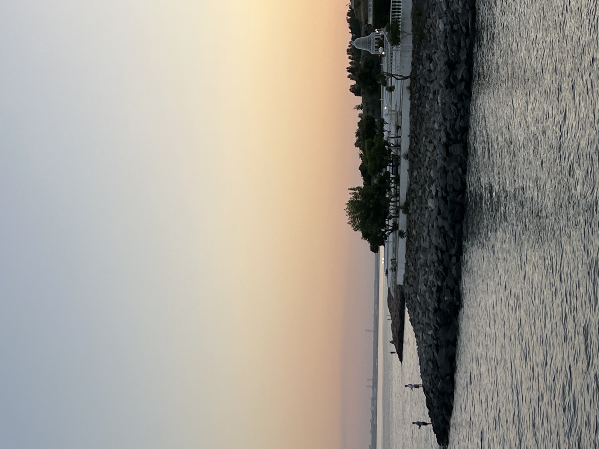 The image depicts a serene coastal scene at sunset in an unspecified location. The sky transitions from a pale blue at the top to hues of pink and orange near the horizon. The sea is calm with gentle ripples reflecting the soft light. In the foreground, a rocky shoreline juts out into the water.
Several individuals are visible on the rocky outcrop, appearing to be fishing. Two figures are more prominent, standing on the rocks with fishing rods. Further along the shore, a few more people are present, some standing and some possibly sitting.
Beyond the rocky area, a paved walkway or terrace is visible, bordered by a white railing. Lush green trees and shrubbery are planted behind the walkway. A distinctive white structure, resembling a small dome or pavilion, stands on the right side of the image. In the distance, across the water, a hazy urban landscape with industrial-looking structures is faintly discernible. The overall atmosphere is tranquil, suggesting an evening by the sea.