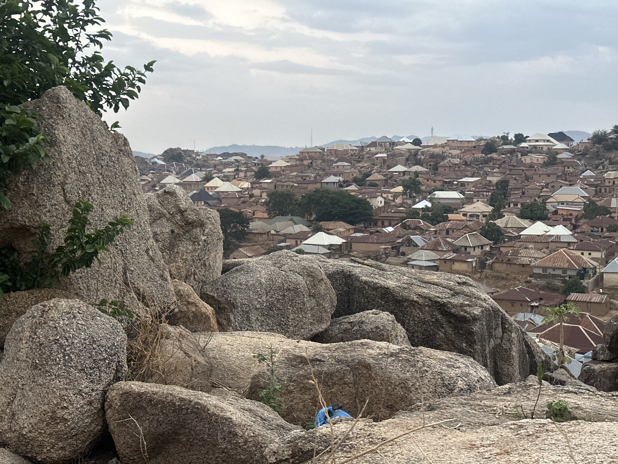 The image shows an overview of a densely populated residential area, likely in Jos, Nigeria, viewed from a high vantage point amidst large, gray boulders. The houses are predominantly single-story structures with corrugated metal roofs, primarily in earth tones, white, and gray. Green trees and shrubs are interspersed amongst the buildings and rocks. The sky is overcast. In the foreground, a cluster of gray rocks with some vegetation partially obscures the view. A small blue object is visible nestled among the rocks. In the distance, the skyline is slightly hilly with faint hints of structures or natural formations.