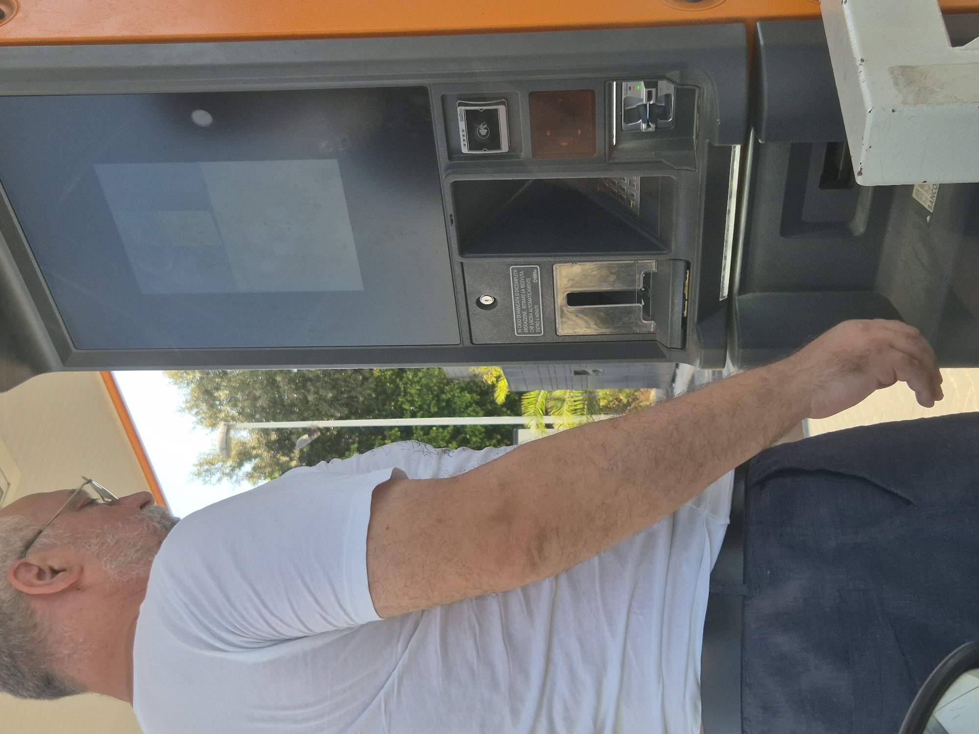 A middle-aged man with glasses and a goatee is standing next to a parking payment machine. He is wearing a white t-shirt and dark pants. The machine has a screen, a card reader, and a slot for coins. There is text on the machine in Italian, indicating instructions for incomplete transactions. The background shows trees and a lamppost, suggesting an outdoor urban setting. The lighting suggests it is daytime. The context implies this is a parking area within the city of Taranto, Italy, likely a public parking facility.