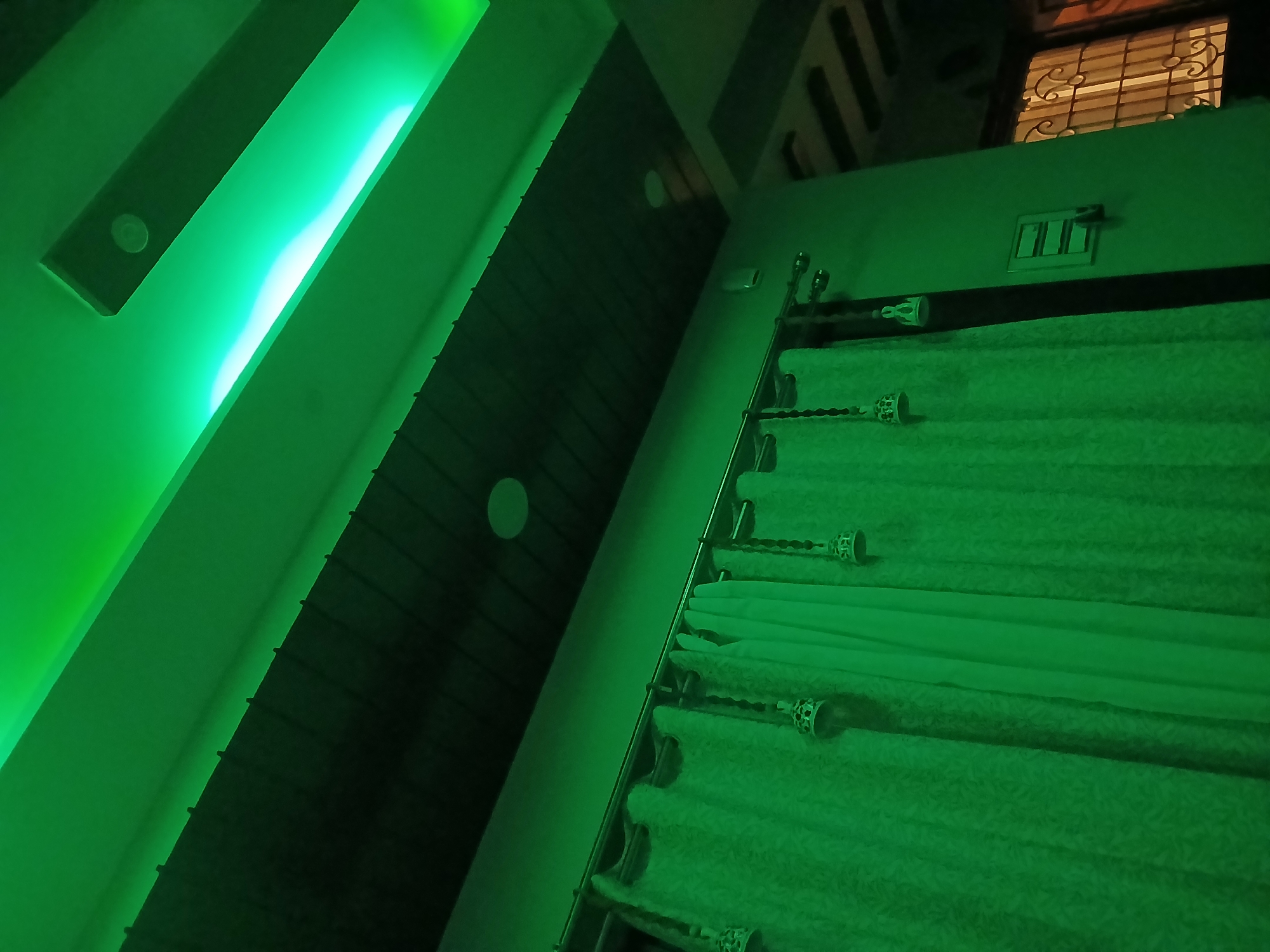 The image captures an indoor room, likely a bedroom or living area, bathed in a strong artificial green light. No people are present, and no specific activity is taking place.
The most prominent feature is a strip of bright green LED lighting installed along a false ceiling or an architectural recess on the left side of the frame, casting its hue across the entire space. Adjacent to this, a dark wooden-paneled or slatted ceiling/wall feature runs diagonally, featuring small circular details, possibly recessed lights or decorative elements.
On the right side of the image, light-colored curtains, appearing green under the ambient lighting, are drawn. They are supported by a metallic curtain rod with decorative finials. Above the curtains, a white rectangular electrical switch plate with multiple switches is mounted on the wall. Partially visible at the top right, a window with a decorative metal security grill can be seen, offering a glimpse of a darker exterior, suggesting it is nighttime.
The overall atmosphere is modern and distinctive, created by the monochromatic green illumination. The room's elements, such as the window grill, align with residential interiors commonly found in Bengaluru, India. No visible text is present in the image.