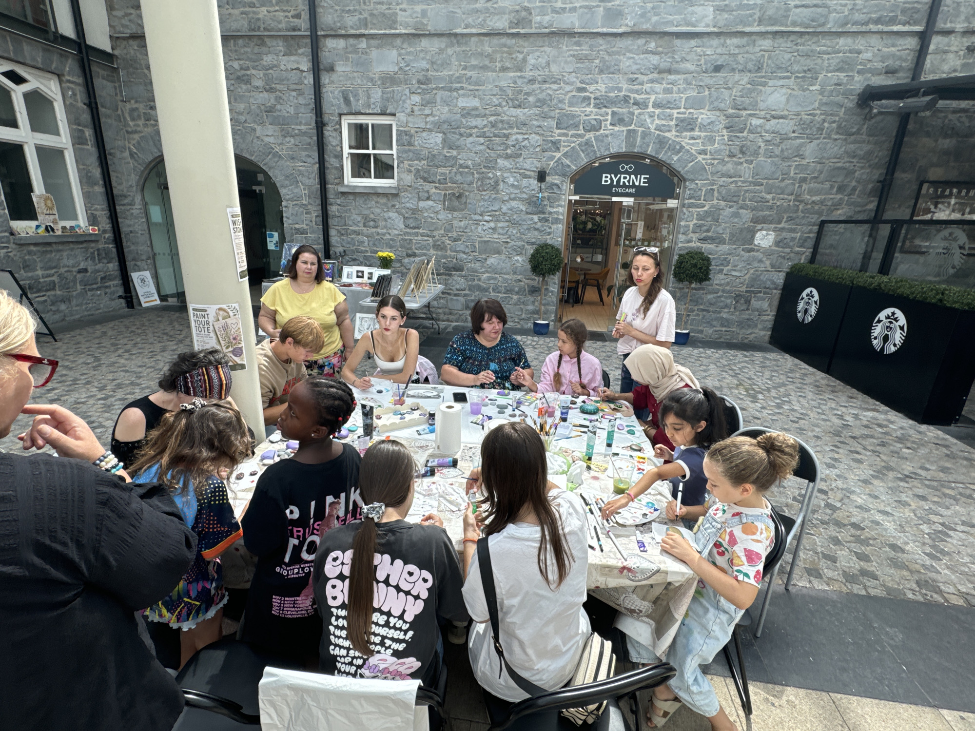 An outdoor arts and crafts workshop is taking place at a long table set up in a courtyard. Several people, mostly children and young adults, are seated around the table, engaged in painting and crafting activities. Art supplies such as paints, brushes, palettes, and paper are spread across the table. In the background, there is a stone building with an arched entrance and signage for "BYRNE EYECARE". Adjacent to this building is a Starbucks coffee shop. The setting appears to be a public space within the city of Kilkenny, Ireland, likely part of a cultural or retail area. The ground is paved with cobblestones, and there are some plants and outdoor seating visible. The overall atmosphere suggests a community event or an educational activity focused on art.